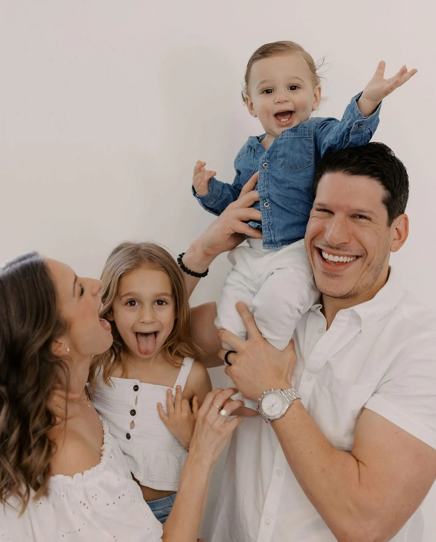 A family of four smiling and playing together against a plain wall, the father holding their toddler son up while the mother and daughter look on.