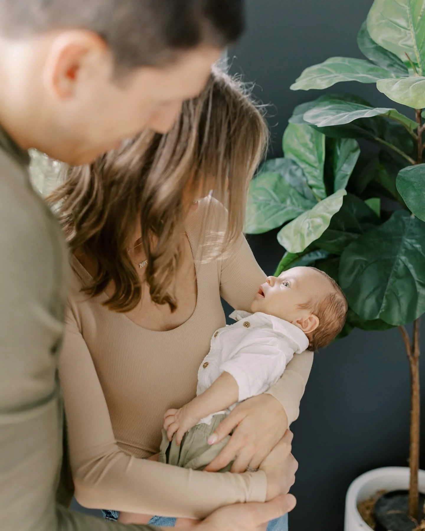 Woman holding a baby while looking at someone out of frame, with a man partially visible next to her, and large green plants in the background.