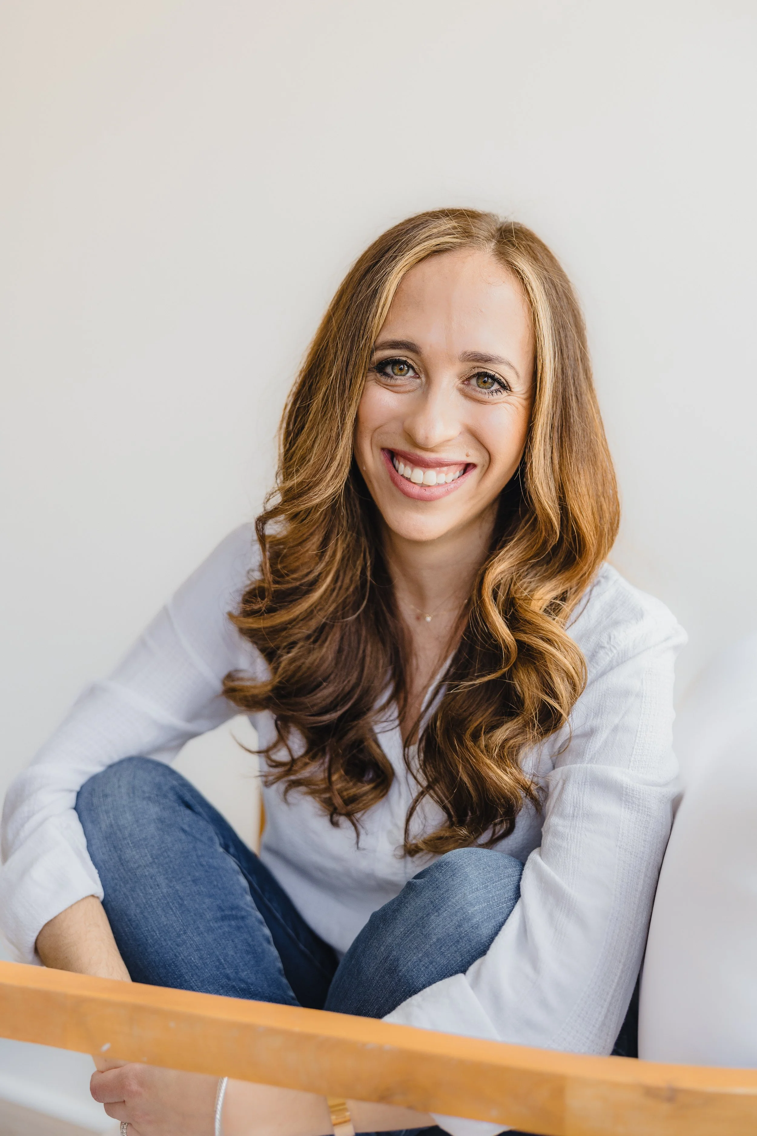 A woman with long, wavy red hair smiling and sitting on the floor with a white background behind her.