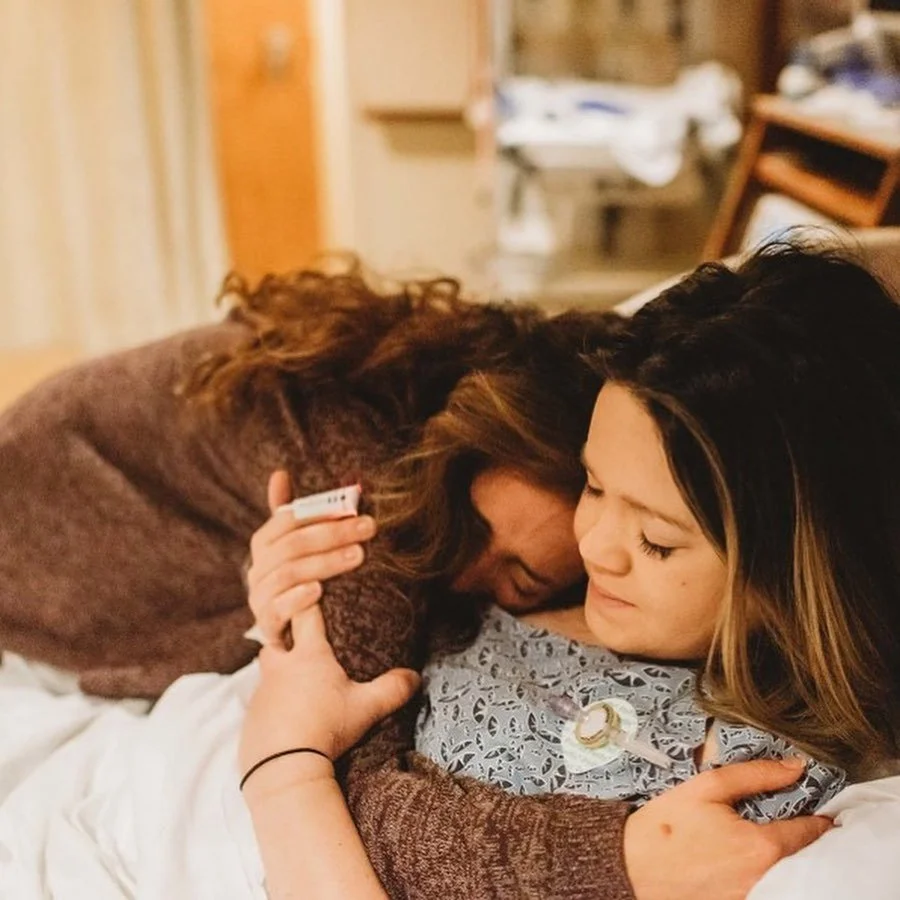 Two women share an emotional embrace in a hospital room, with one woman in a hospital gown lying in bed and the other woman kneeling beside her.