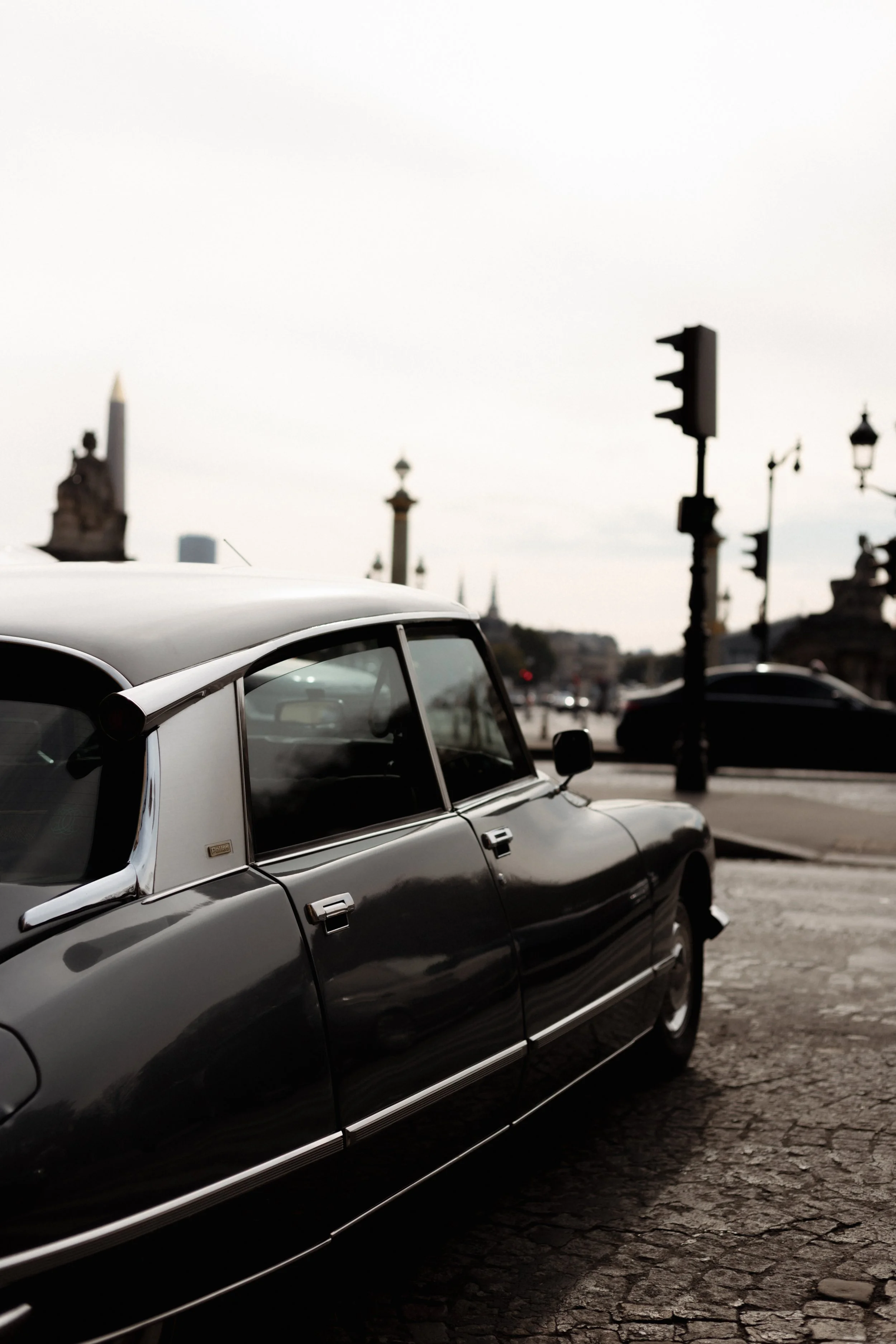 A vintage black car parked on a cobblestone street in front of streetlights and historical monuments, with a cloudy sky overhead.