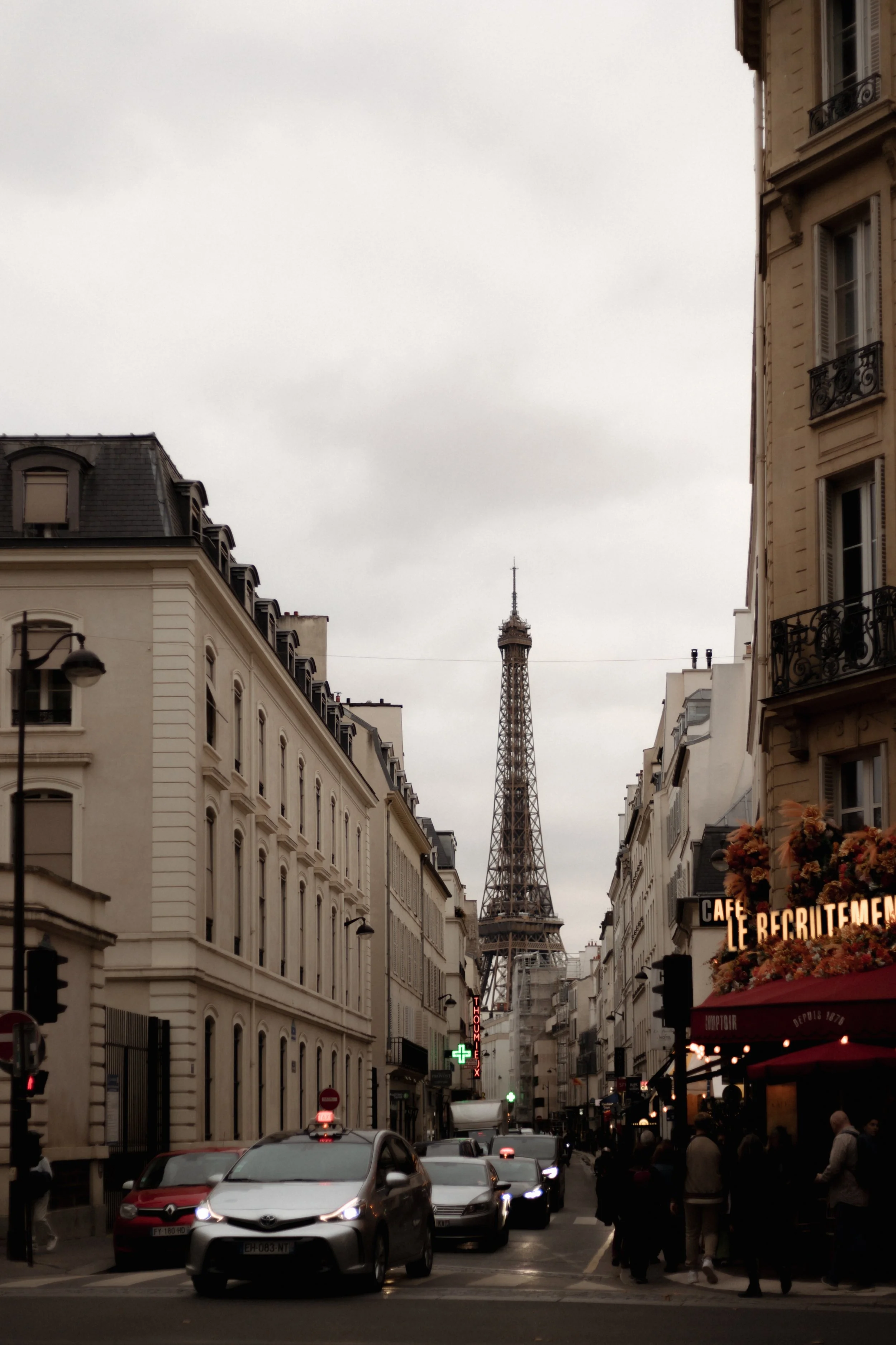 Street view in Paris with cars on the road, pedestrians on the sidewalk, buildings on either side with wrought-iron balconies, and the Eiffel Tower in the distance under a cloudy sky.