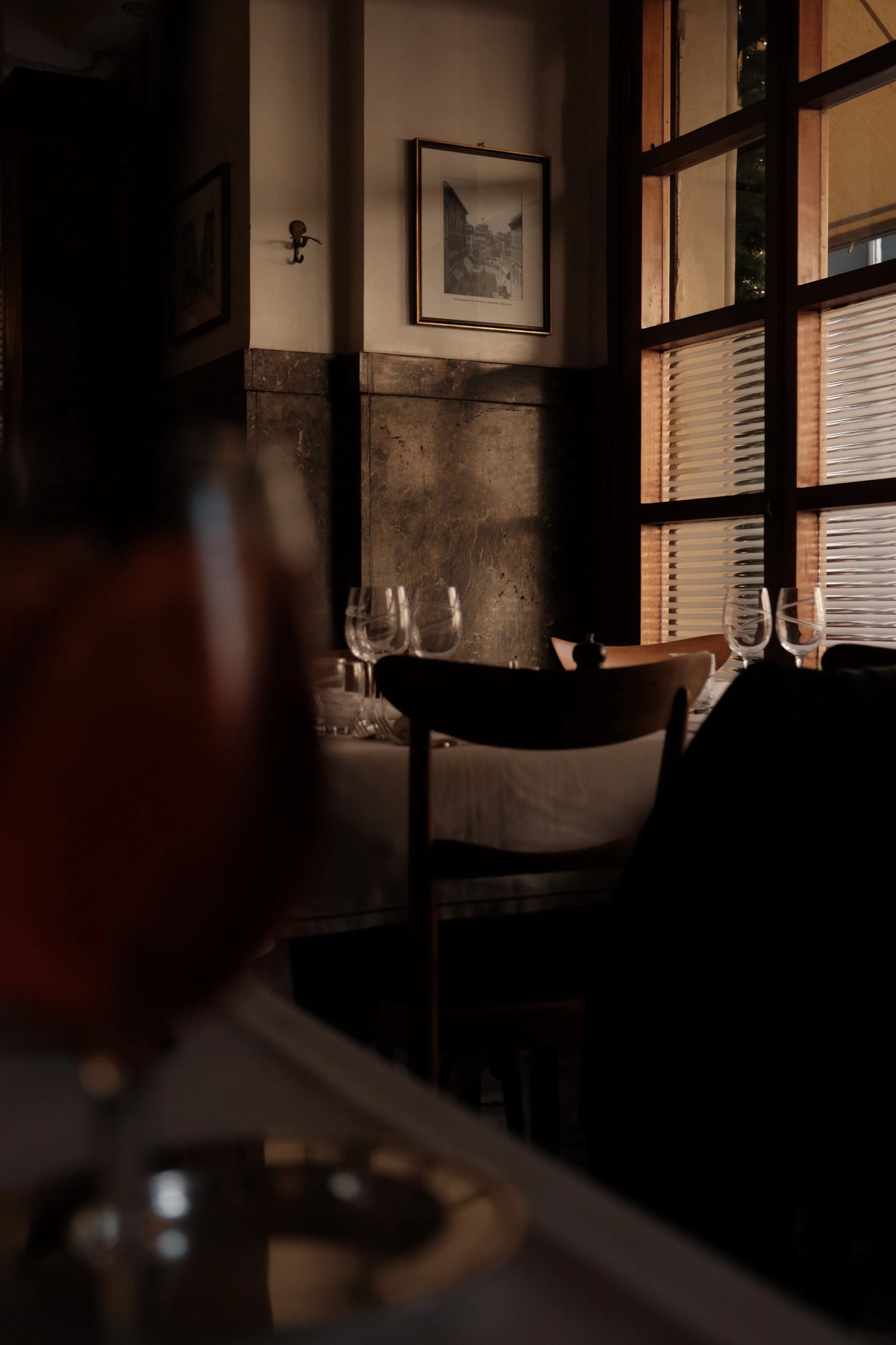 Dimly lit restaurant interior with framed artwork on the wall, wooden window frames, and a table set with wine glasses and a bowl.