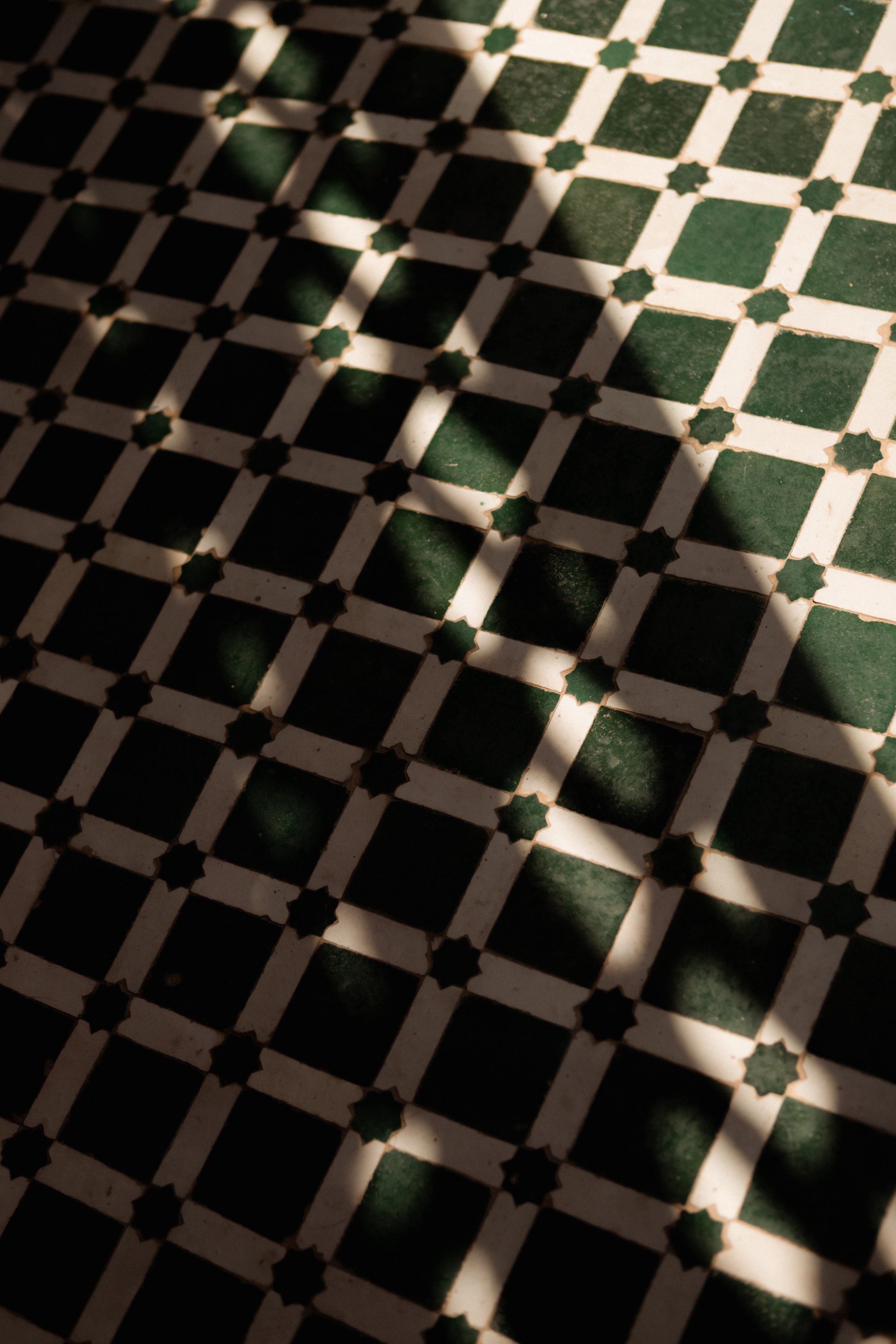 Close-up of a patterned tiled floor with black and white tiles arranged in a geometric pattern, partially covered by shadows from an object or structure outside the frame.