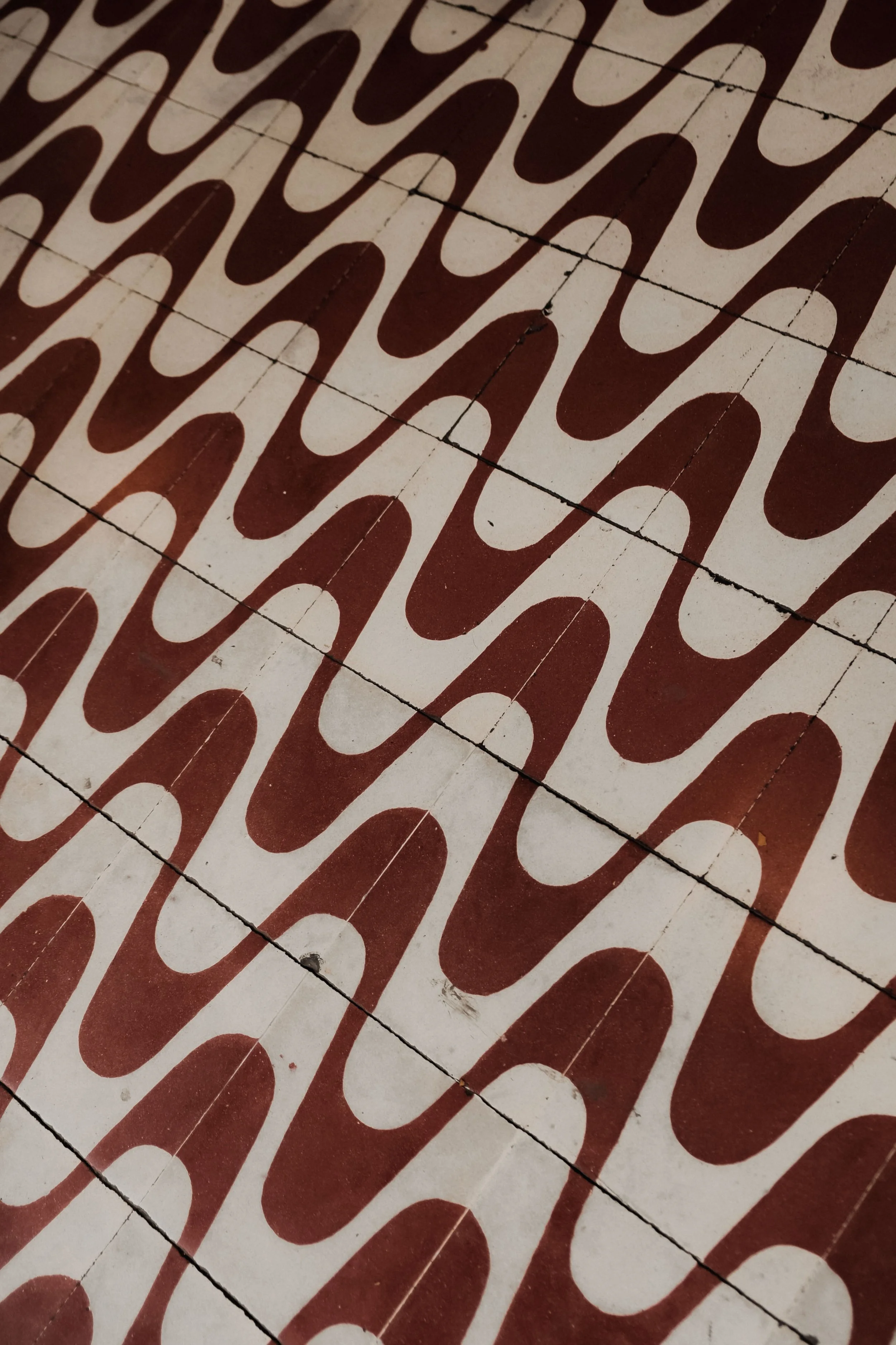 Square ceramic tiles with a pattern of large, brown, elongated loops on a cream background, arranged in a grid with visible black grout lines.