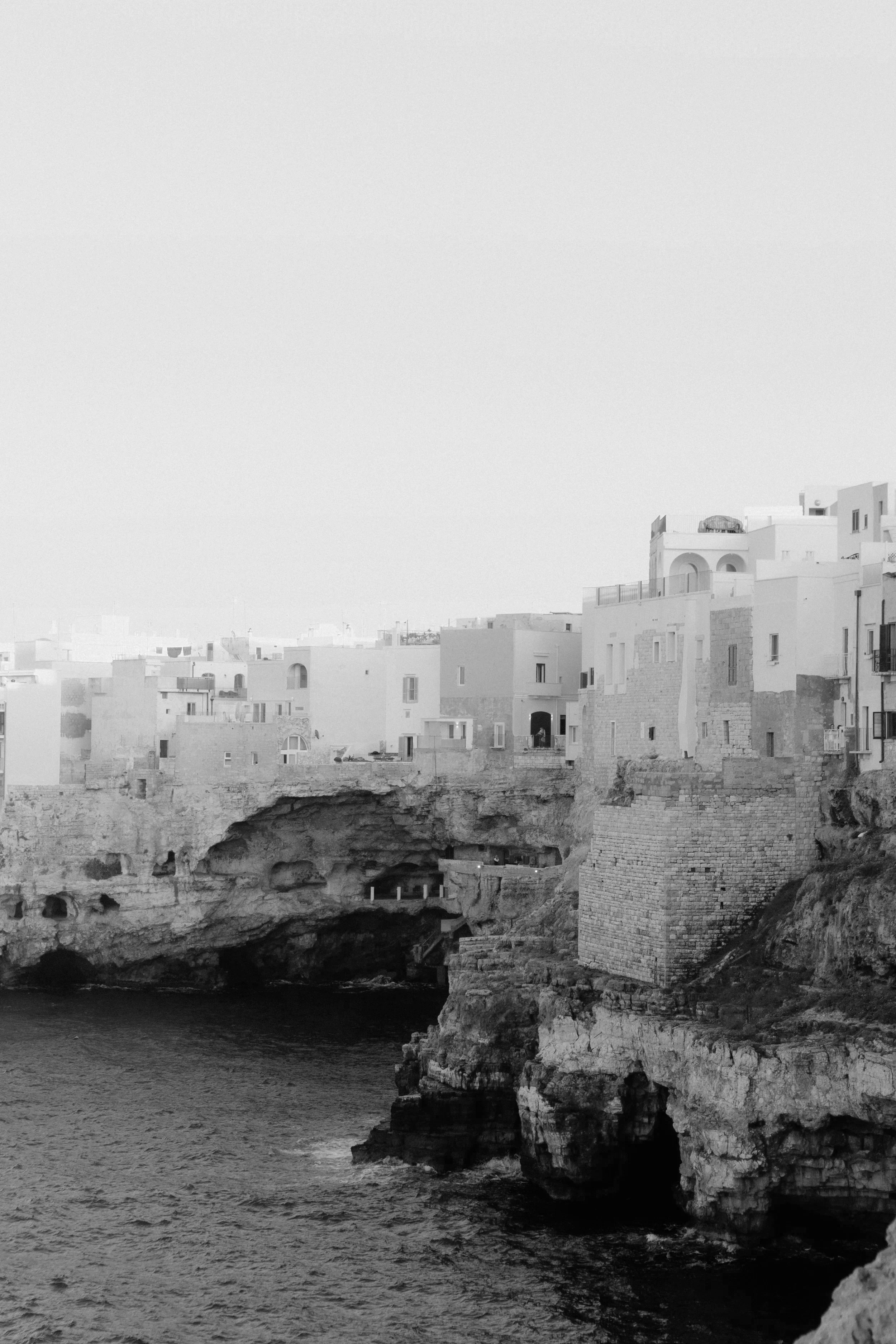 Black and white photo of coastal buildings perched on cliffs above the ocean, with rocky caves at the water's edge.