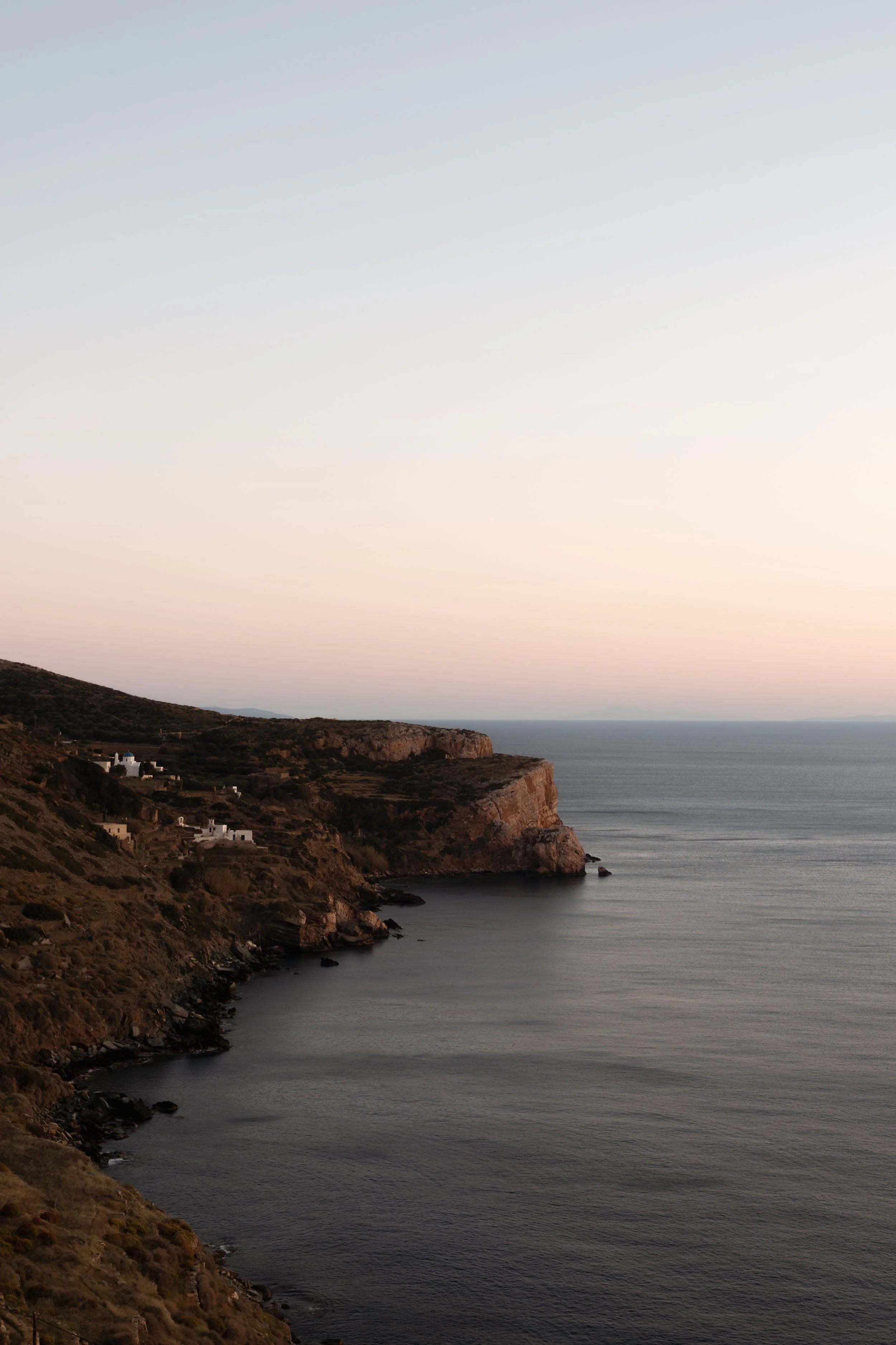 Coastal landscape at sunset with cliffs, scattered houses, and calm ocean water.