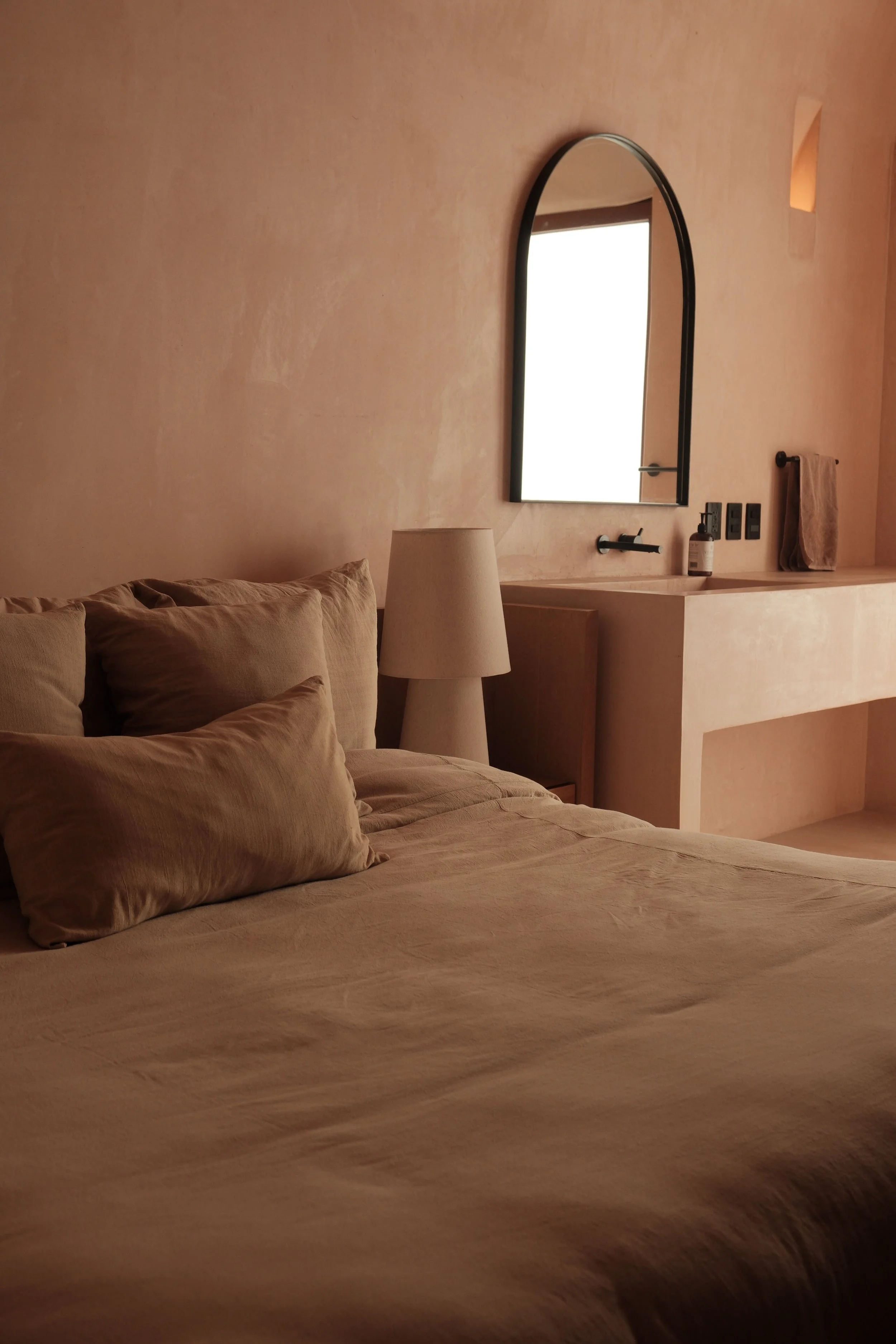 A cozy bedroom with beige bedding and pillows, a bedside lamp, and a mirror on the wall, with a towel and a bottle on the counter.