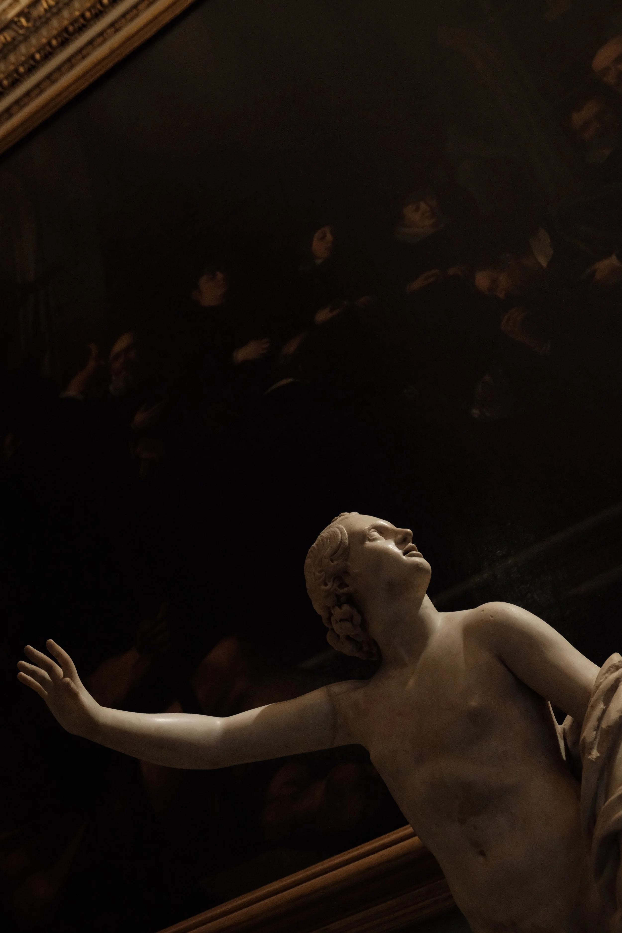 A white marble statue of a woman with her arm extended outward, set against a dark background with reflection of people and books overhead.
