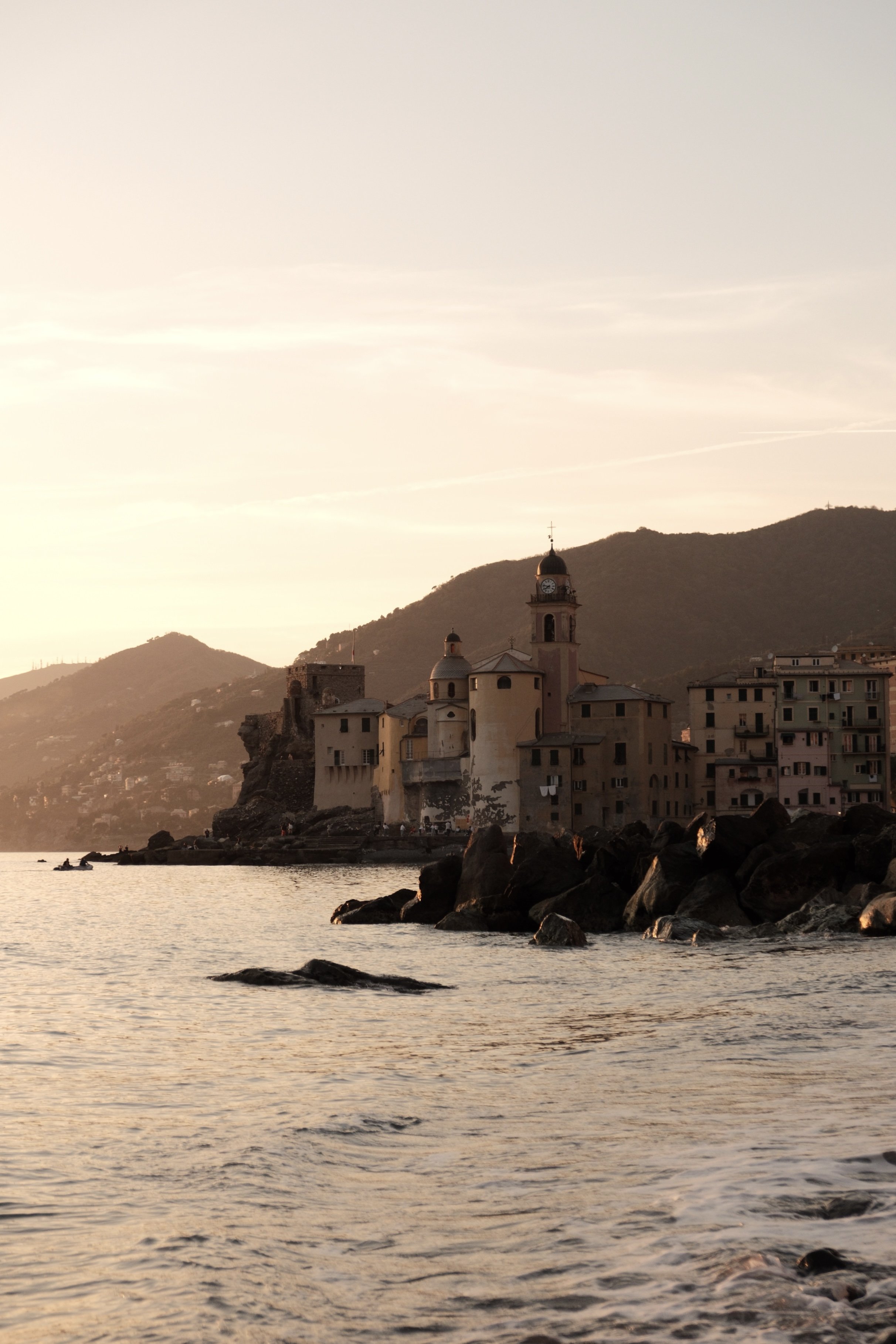 A coastal scene at sunset showing a small town with a church featuring a clock tower, surrounded by mountains and rocks along the shoreline, with calm water in the foreground.