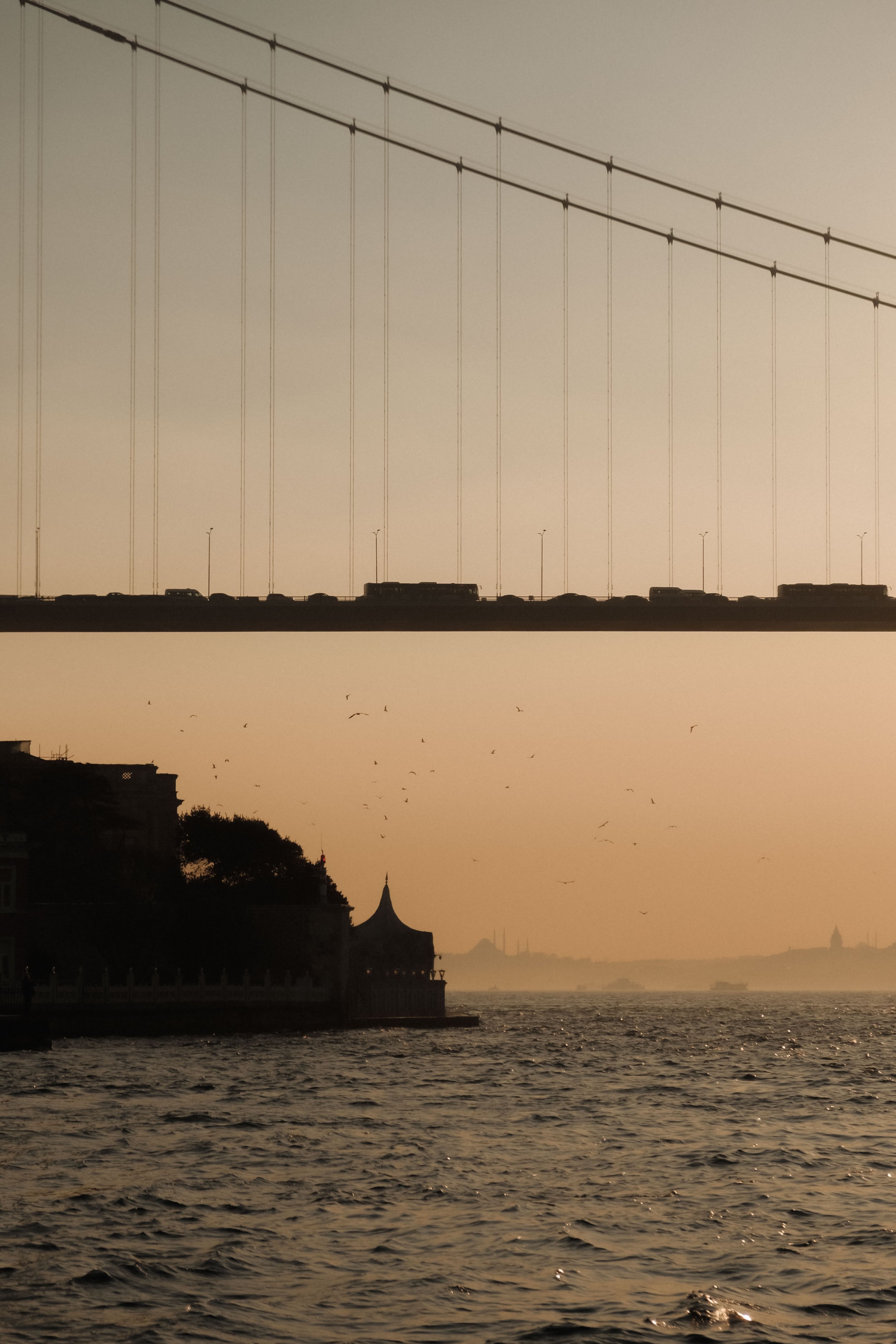 A view of a suspension bridge over water during sunset or sunrise, with the bridge's cables and vehicles visible, and a silhouette of buildings and a church steeple along the shoreline.