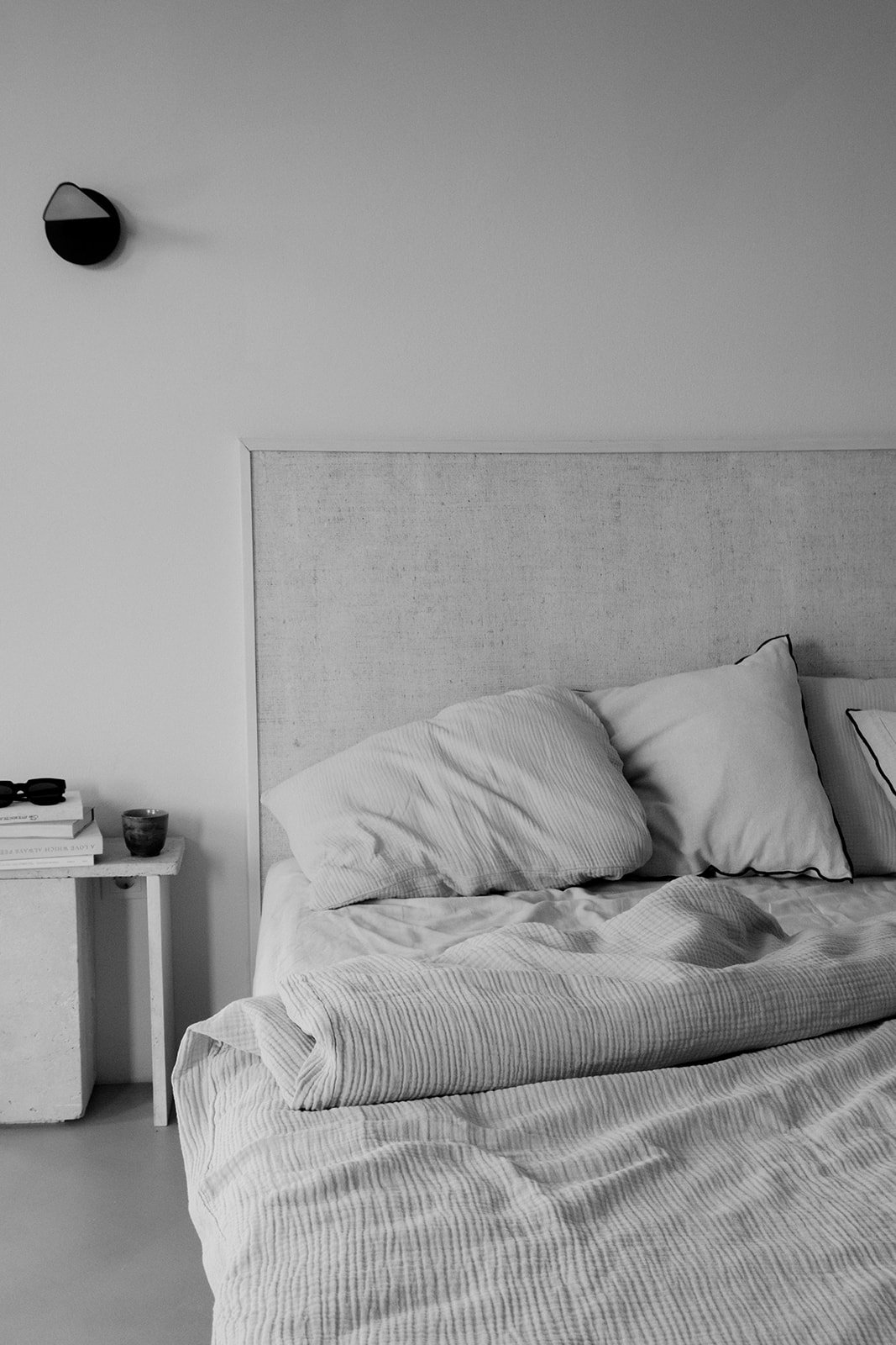 A black and white photo of a minimalist bedroom with unmade bed, including two pillows, a linen bedsheet, a textured headboard, a small white nightstand with books, a cup, and a pair of glasses, and a wall-mounted light fixture on the wall to the left.