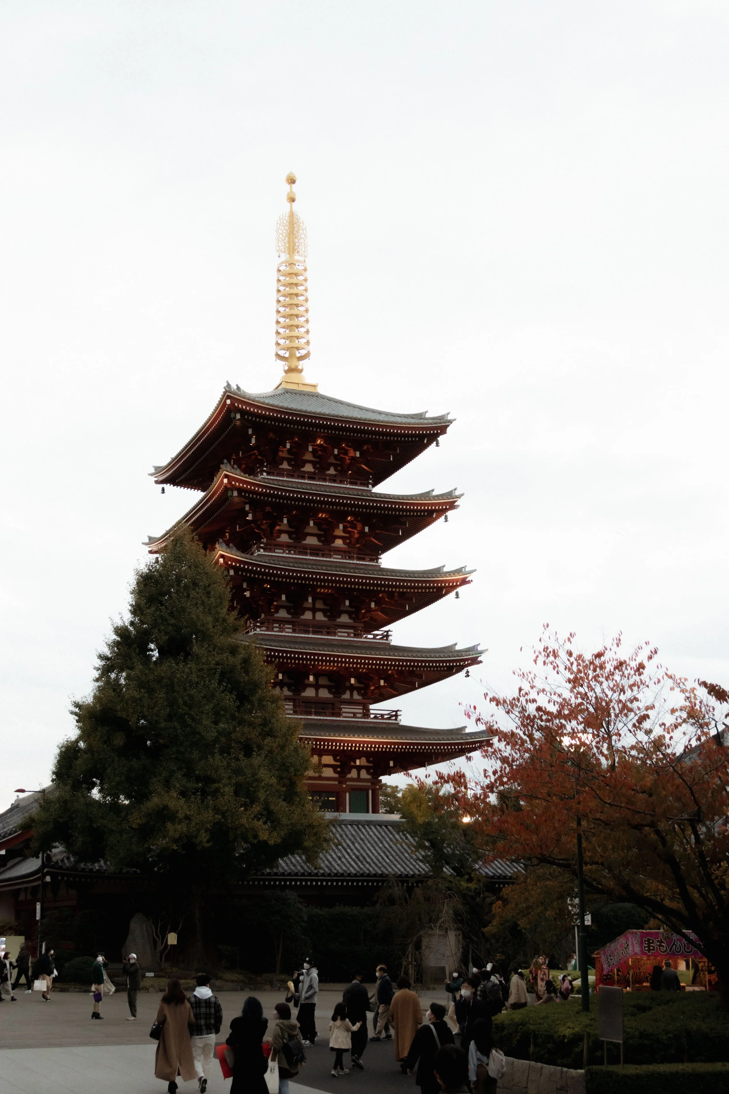 People gather in front of a traditional five-story pagoda in Japan, surrounded by autumn foliage, overcast sky.