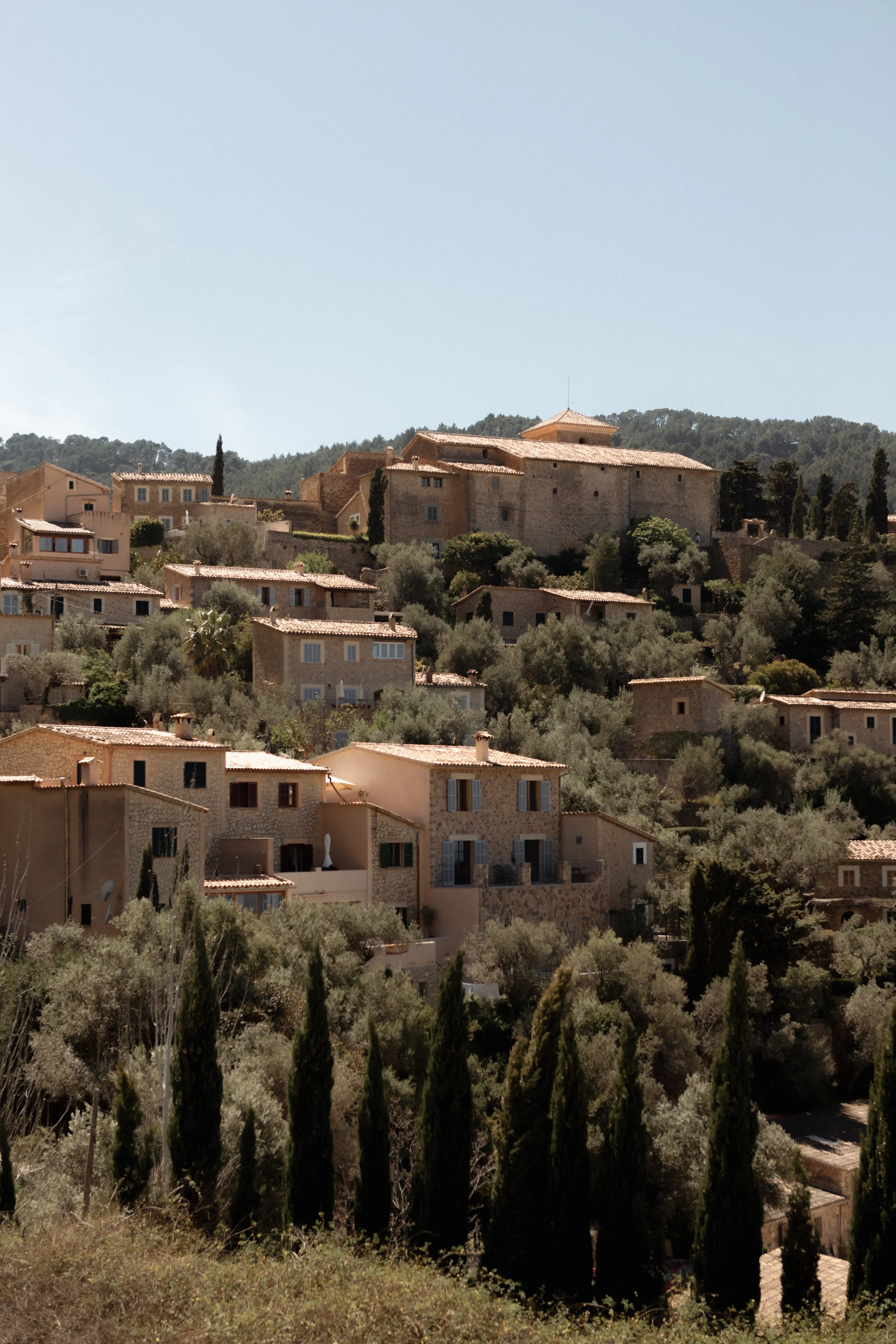 Hilly landscape with stone buildings and Mediterranean-style houses, surrounded by trees and shrubs under a clear blue sky.