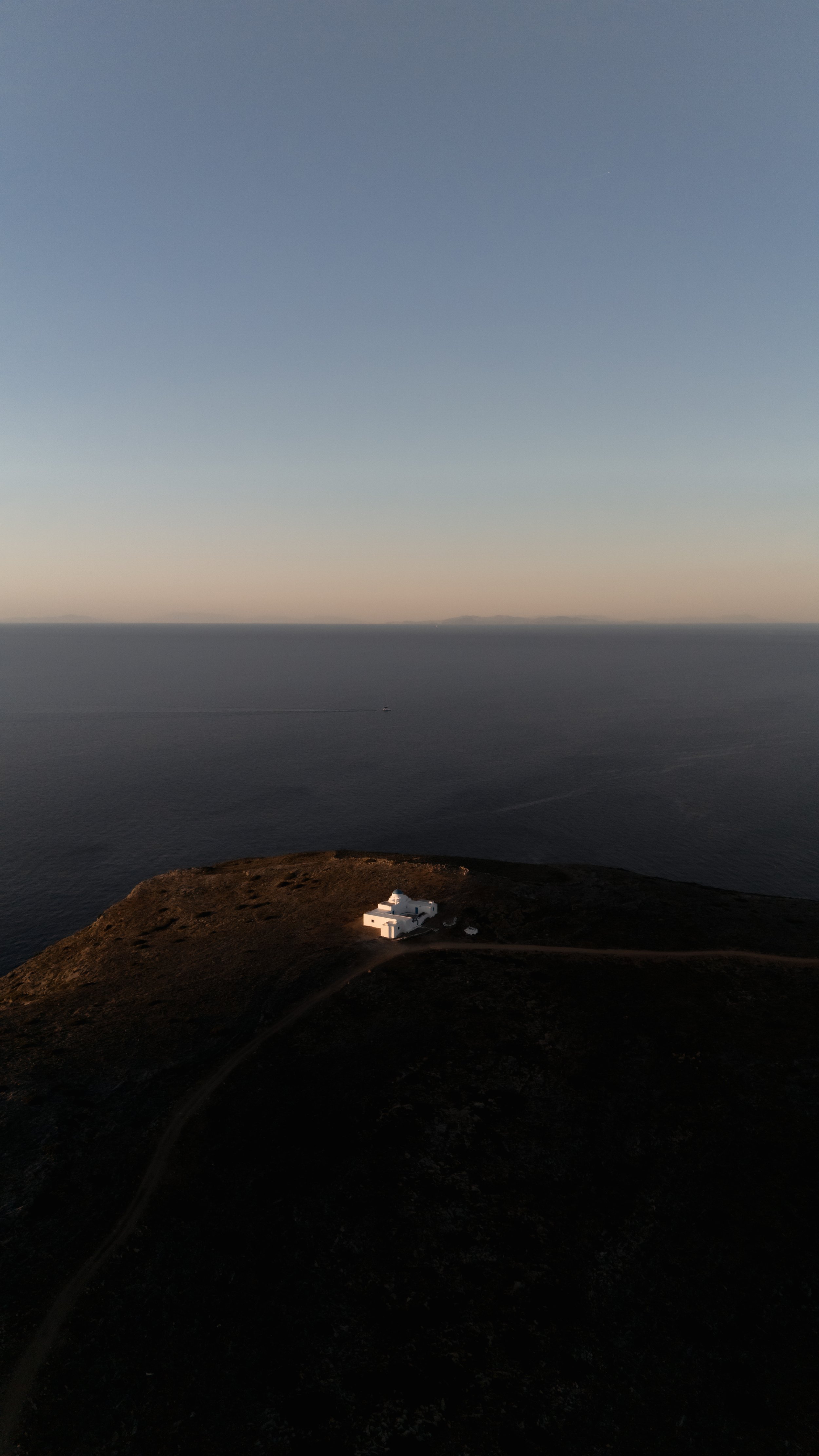 A white building on a hill overlooking the ocean with a small boat in the water and land in the background at sunset or sunrise.