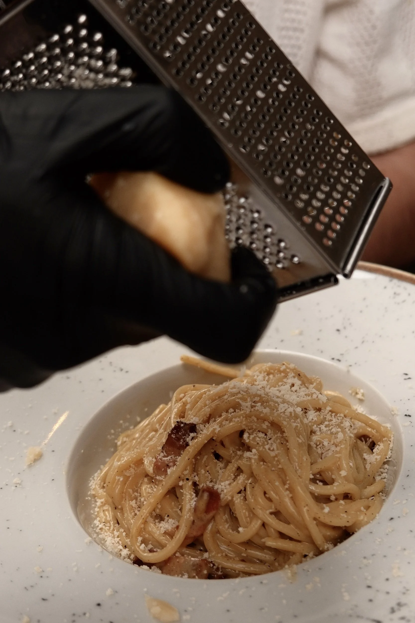 Close-up of a plate of spaghetti pasta with grated cheese, with a hand grating cheese on top, and a metal cheese grater in the background.