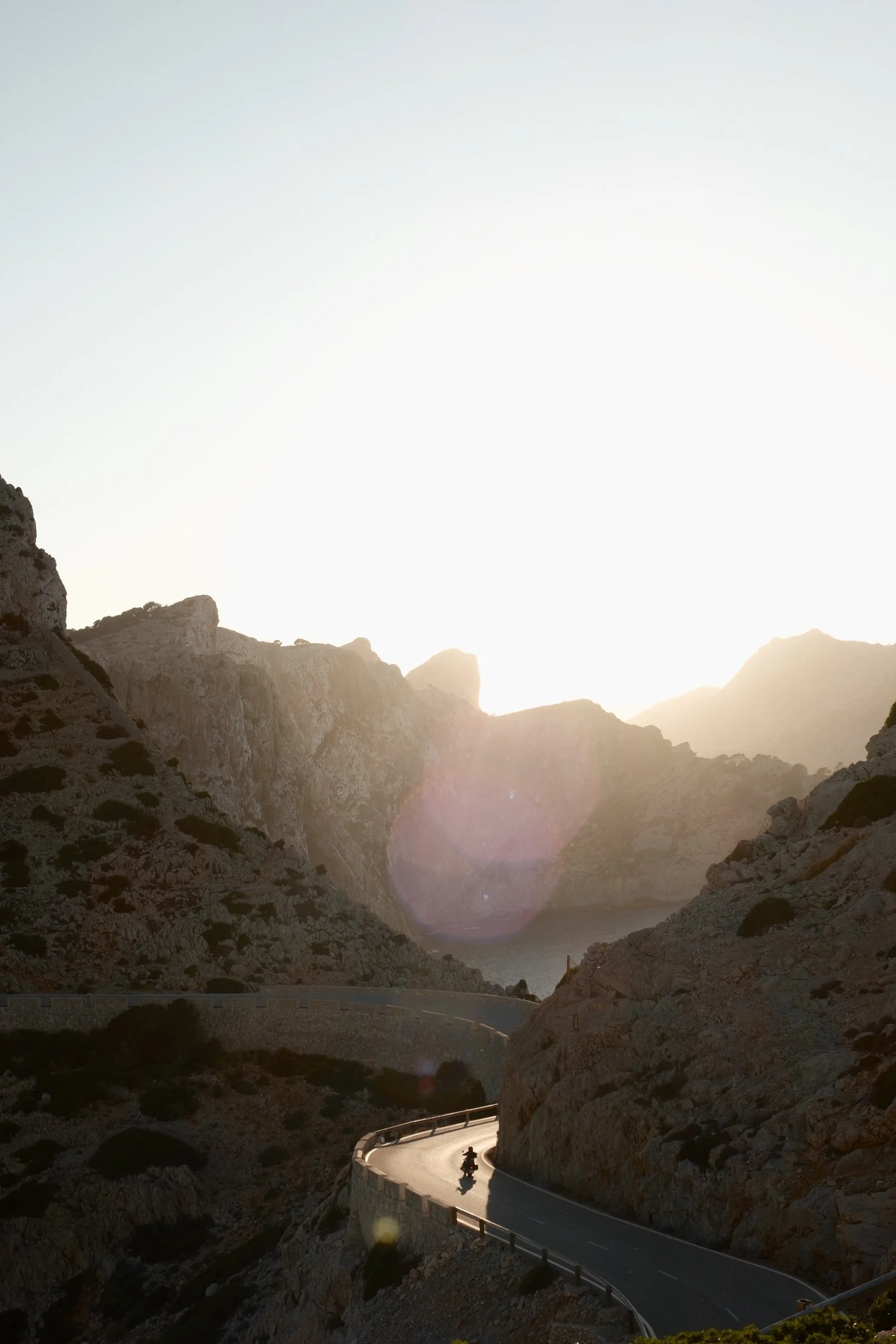Sunset over rugged mountains with a winding road in the foreground, a motorcycle traveling along it, and a distant body of water at the base of the mountains.