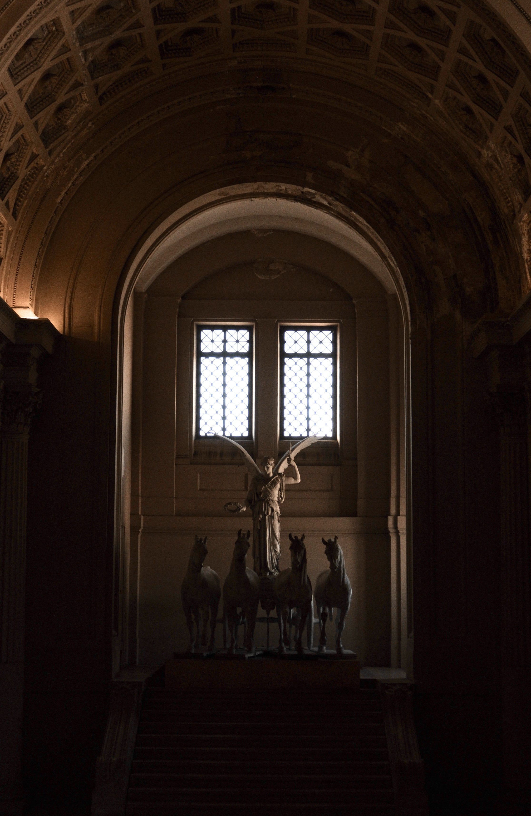 A dimly lit hall with arched ceiling and detailed architecture, featuring a statue of a woman with wings and a crown, surrounded by six horses, and a window at the back letting in natural light.