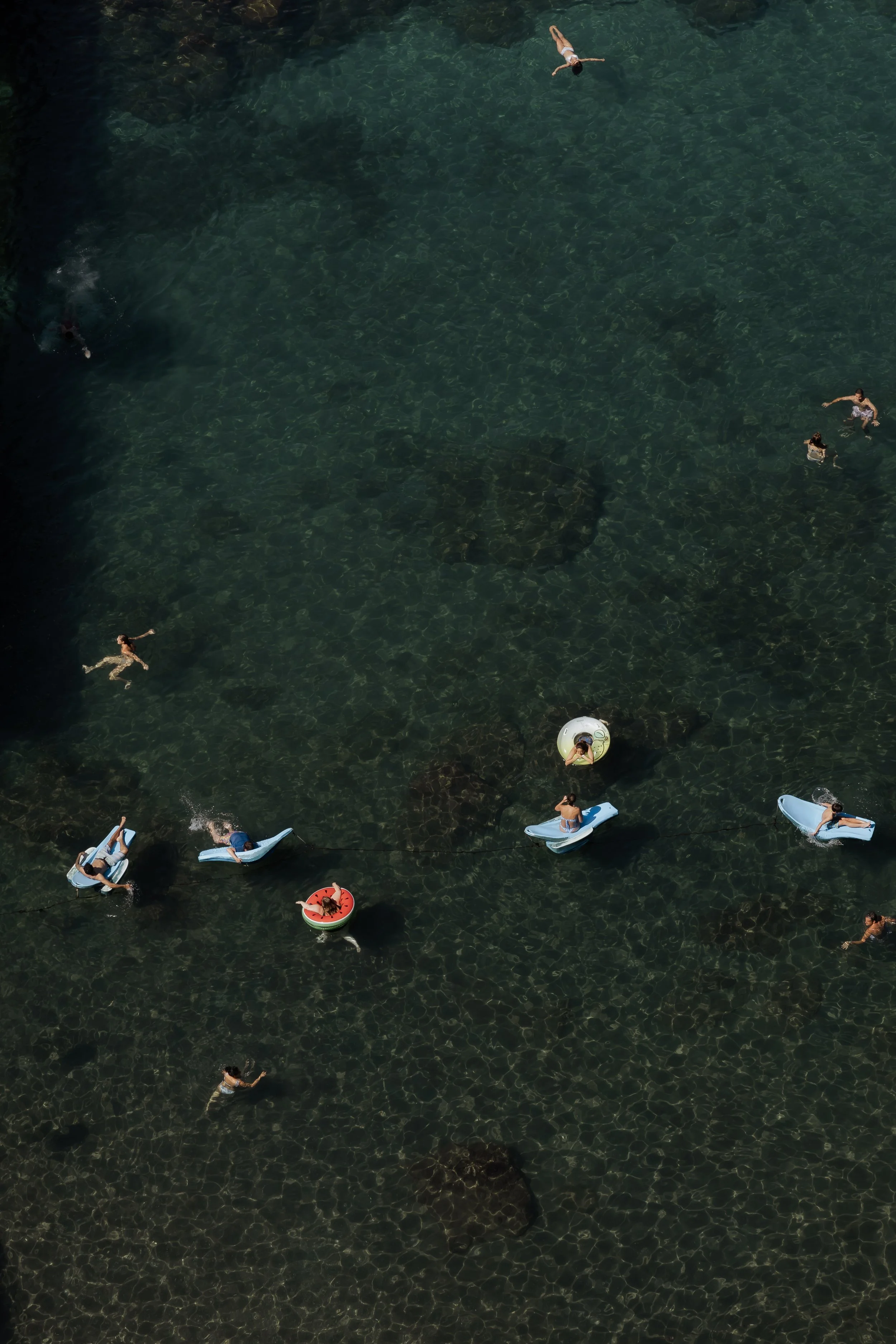 An aerial view of people swimming and relaxing in a clear, greenish water body, some on small boats and a flotation ring.
