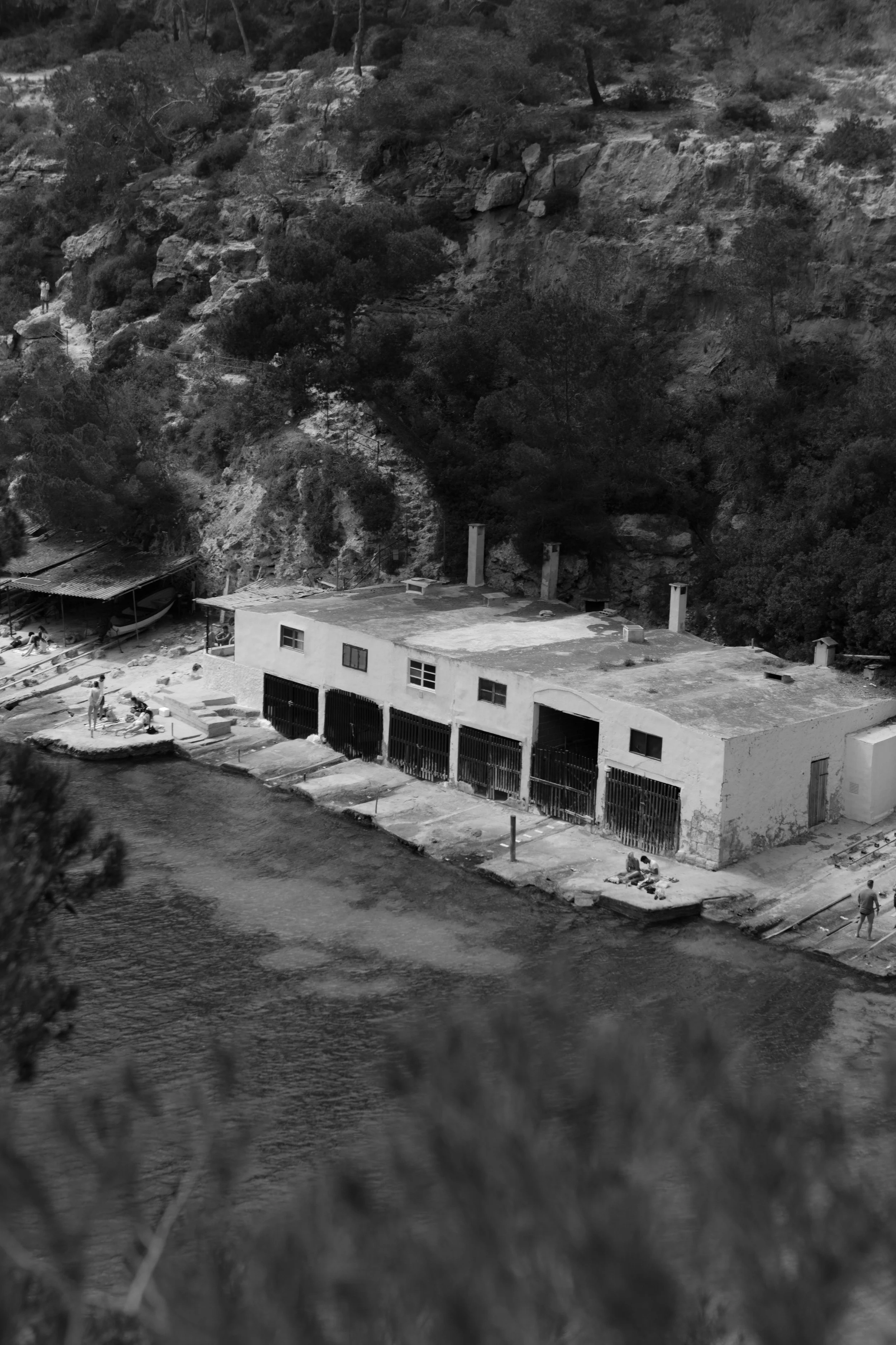Black and white photograph of a building near a body of water with trees and rocky hills in the background, taken from a slightly tilted angle.