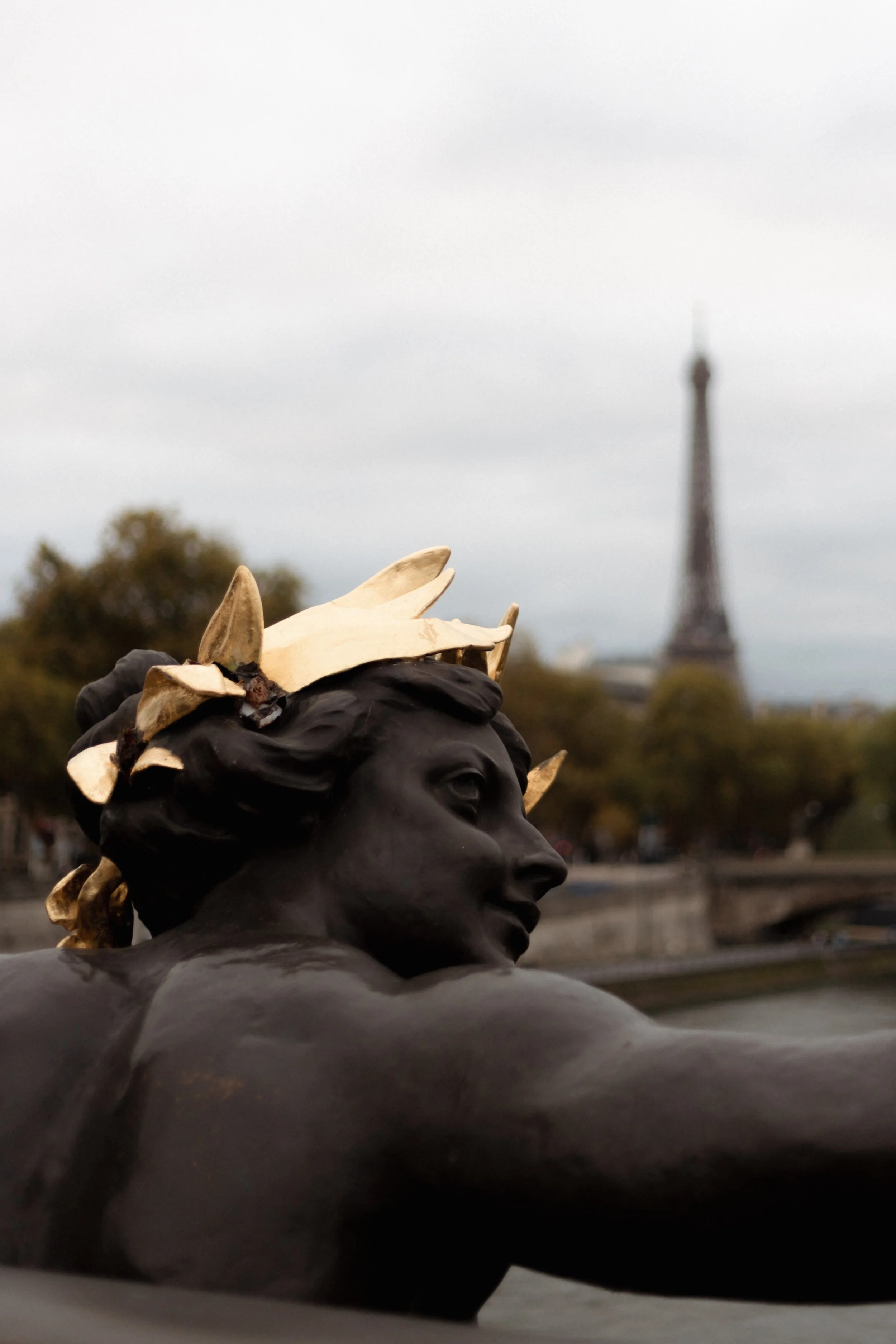 Close-up of a sculpture of a person's head with a crown made of bones, with the Eiffel Tower visible in the background on an overcast day.