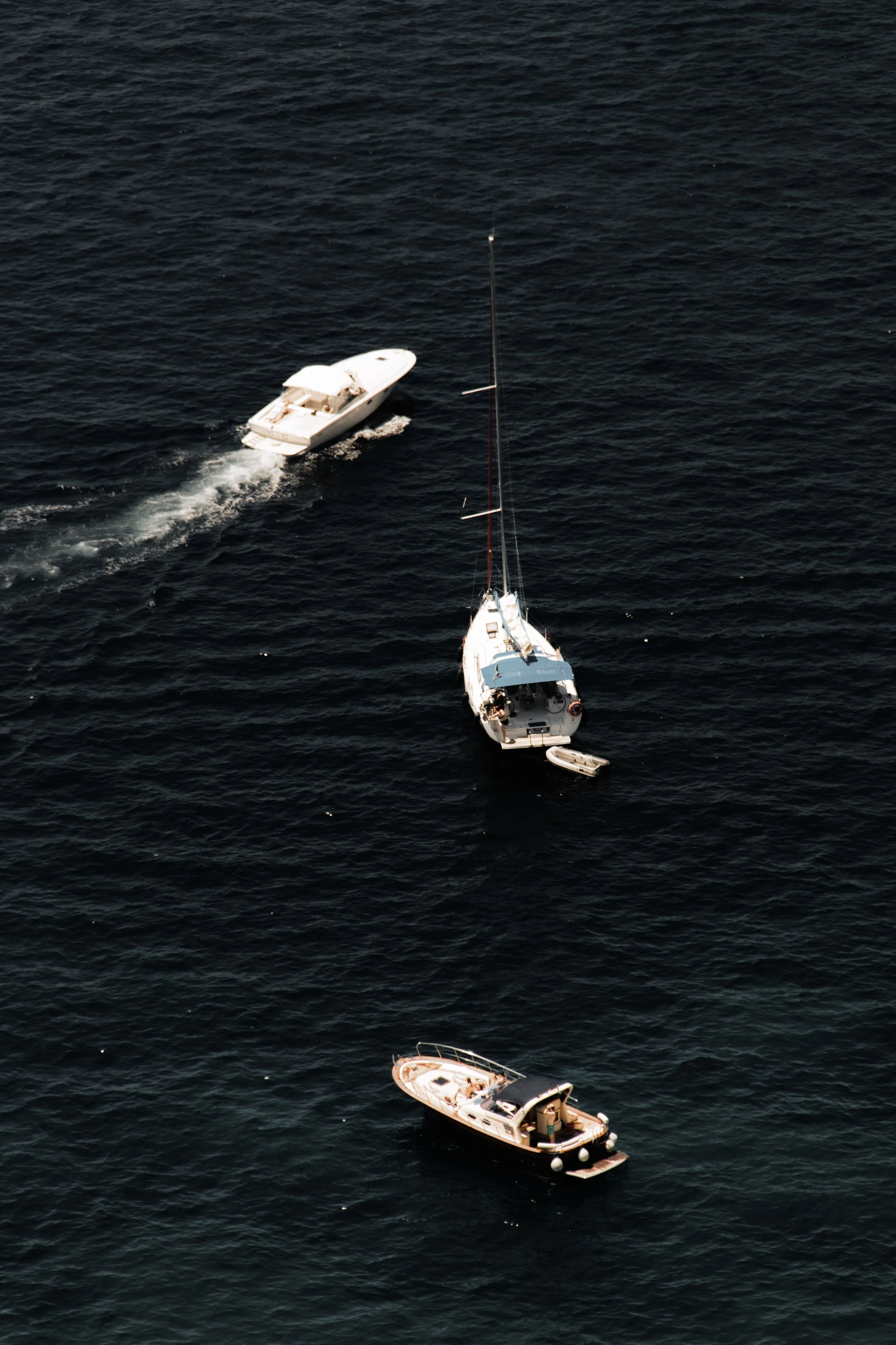 Aerial view of three boats sailing on dark ocean water, one in the foreground with a person on deck and a smaller boat attached, and two other boats further out.