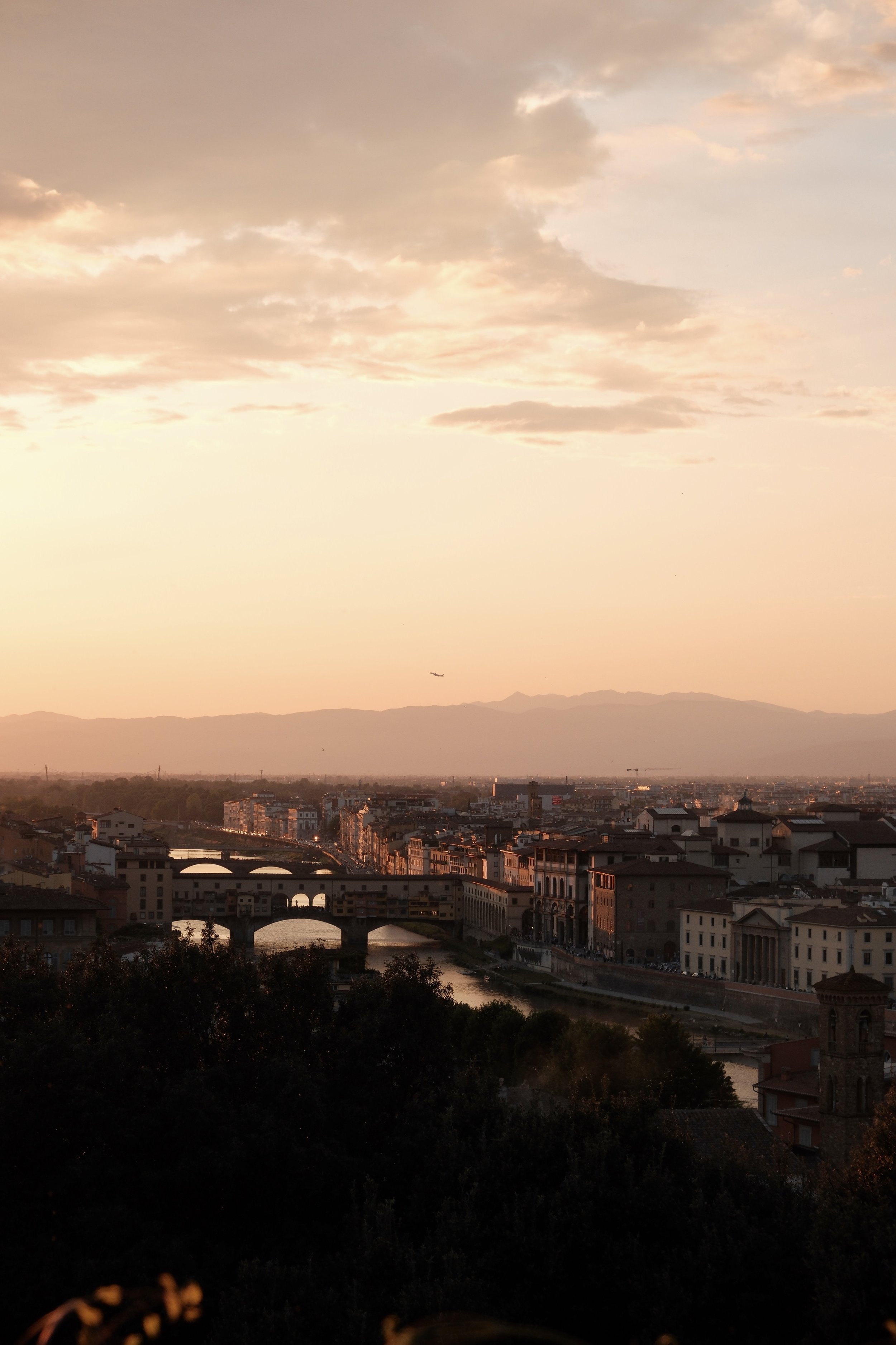 A cityscape at sunset with historic bridges crossing a river, surrounded by low-rise buildings and mountains in the distance, under a sky with clouds and a plane flying.