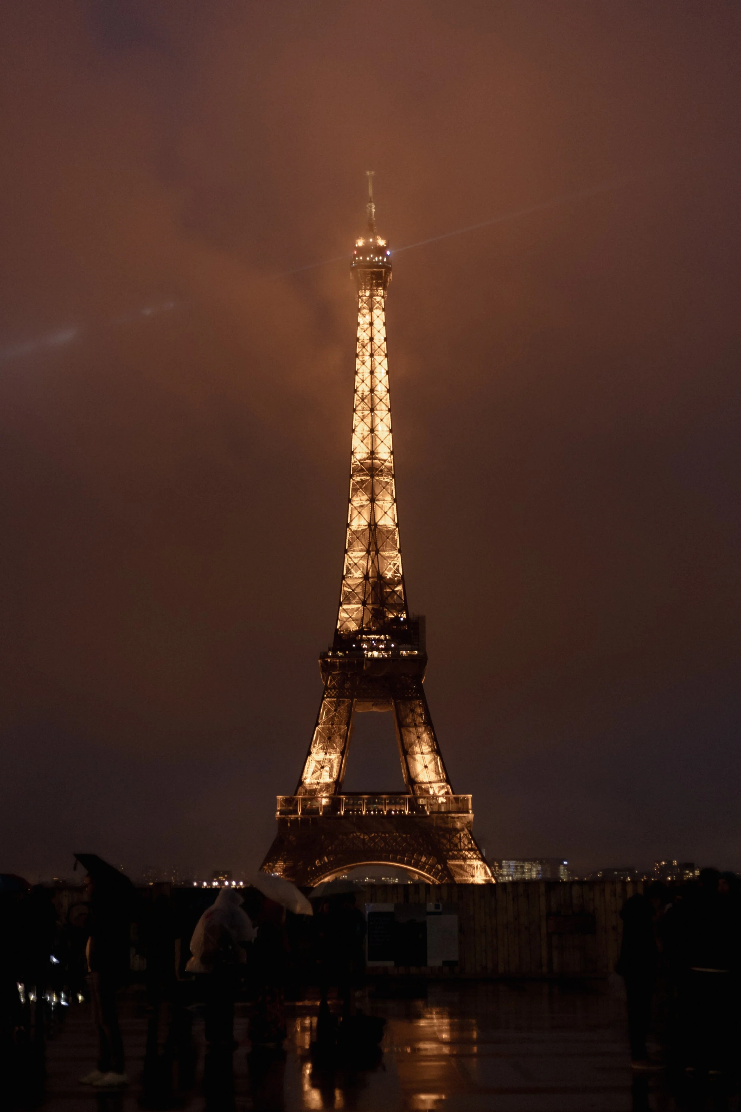 The Eiffel Tower illuminated at night, with people standing in front observing it. The sky is dark and filled with clouds.