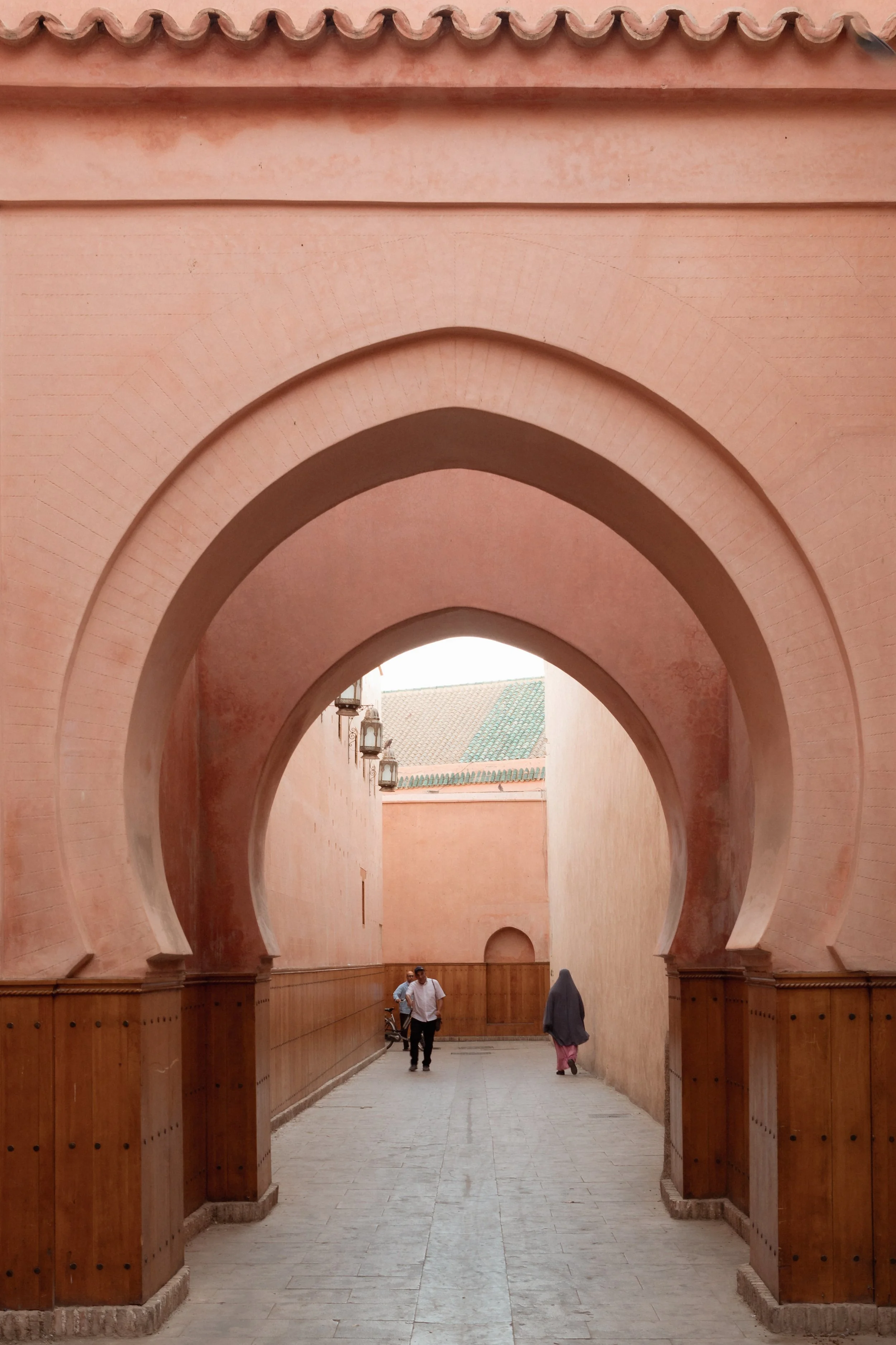 A corridor with pinkish walls and arches, featuring traditional lanterns and two people walking, possibly in an old city or historical site.