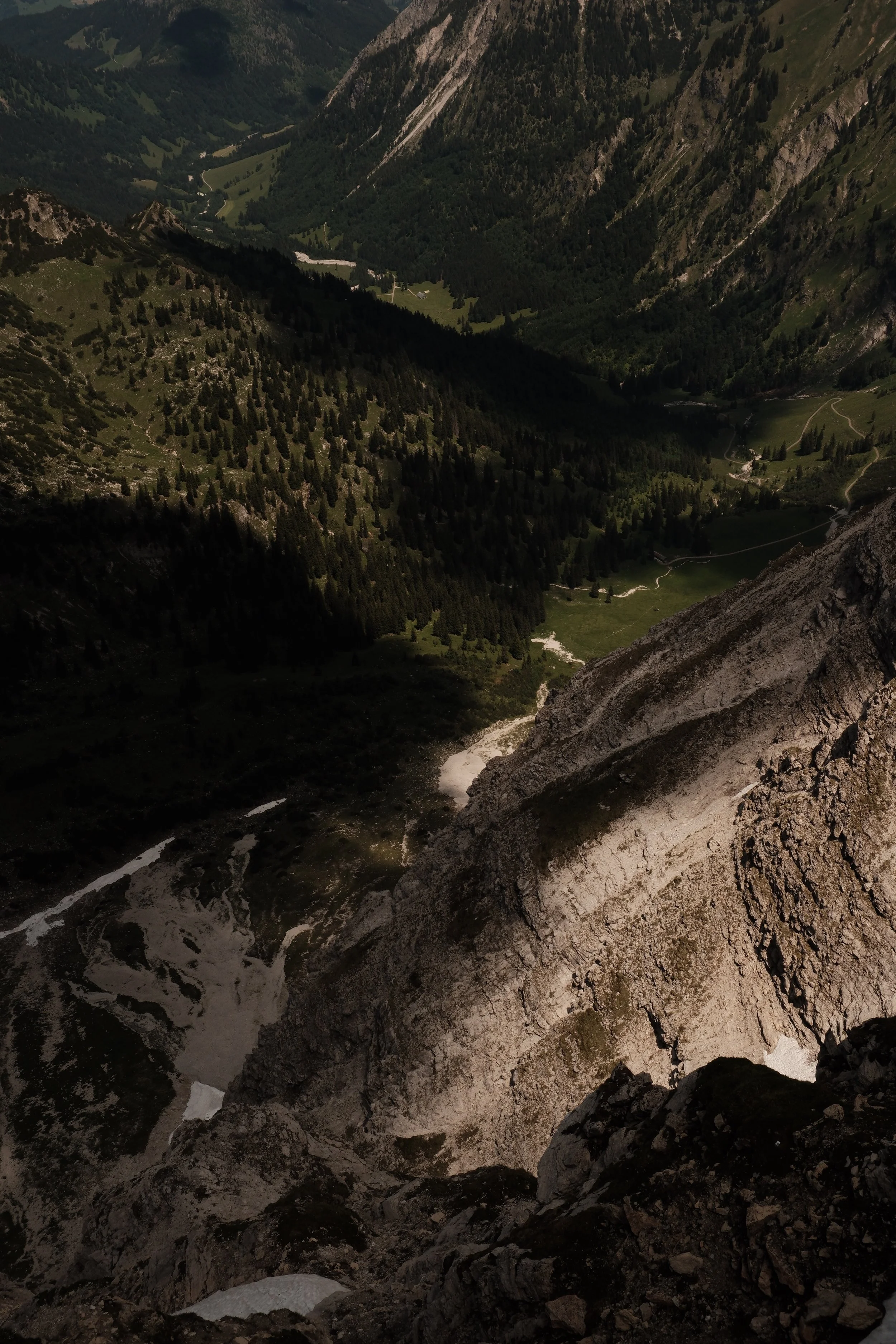 View down into a mountain valley with a small waterfall flowing over rocks, surrounded by steep rocky cliffs and patches of snow, with distant lush green valleys and forested slopes.