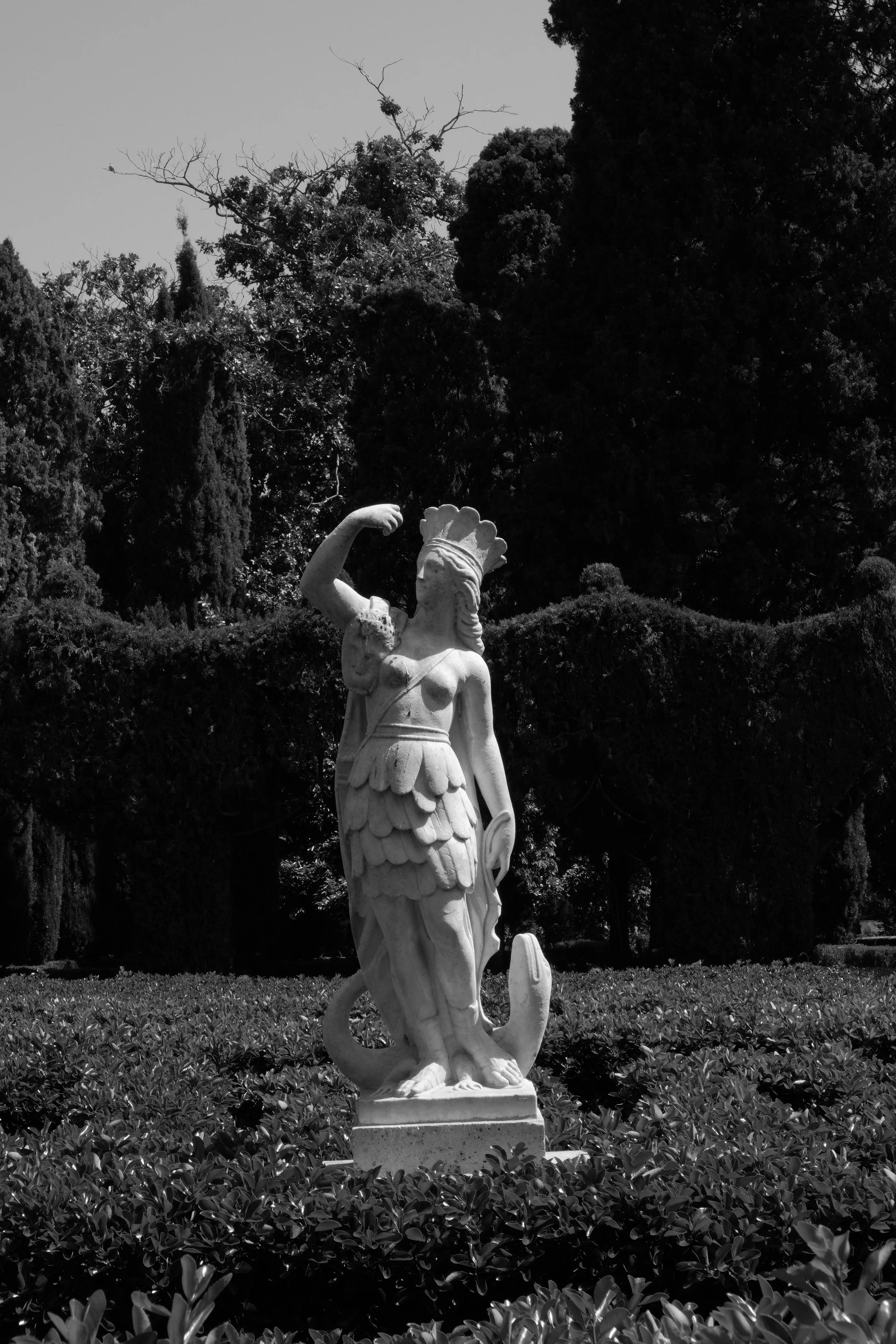 A white marble sculpture of a woman in a garden surrounded by bushes, trees, and cloudy sky.