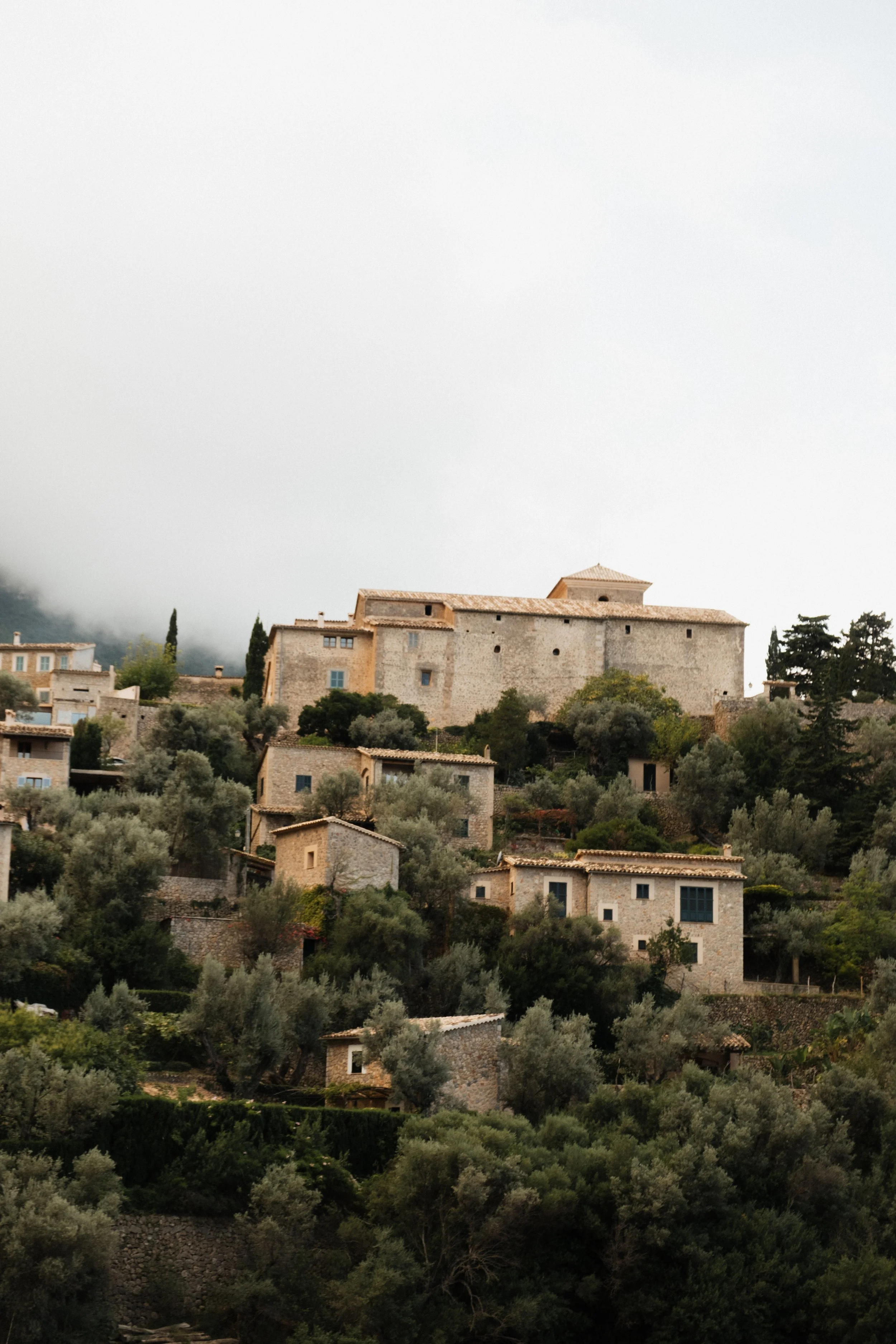 Hilly landscape with stone houses, trees, and a large historic building on a foggy day.