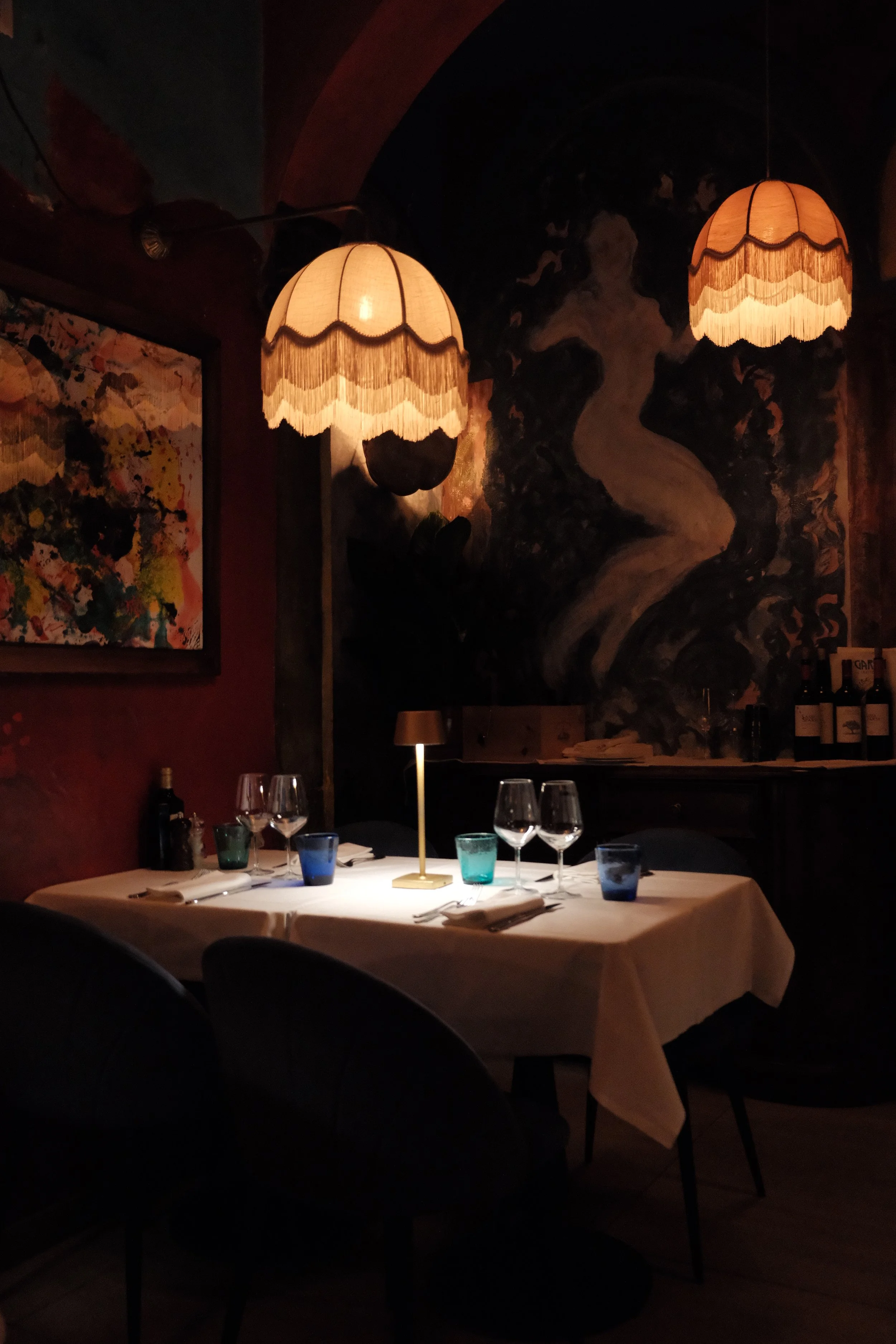 A dimly lit restaurant table set for dining with a white tablecloth, wine glasses, water glasses, napkins, and silverware. Two vintage-style lamps with fringed shades illuminate the table. The background features abstract artwork on the wall and bottles of wine on a counter.