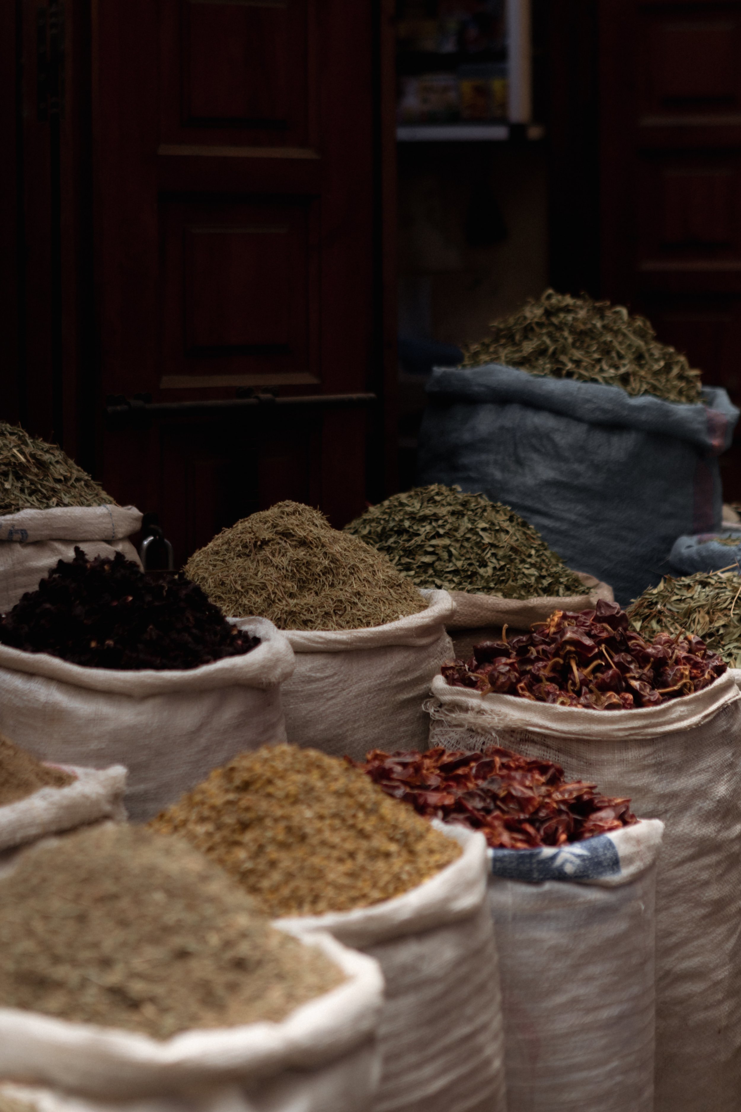 Bags filled with various dried herbs, spices, and teas on a table in a market or store, with wooden cabinets in the background.