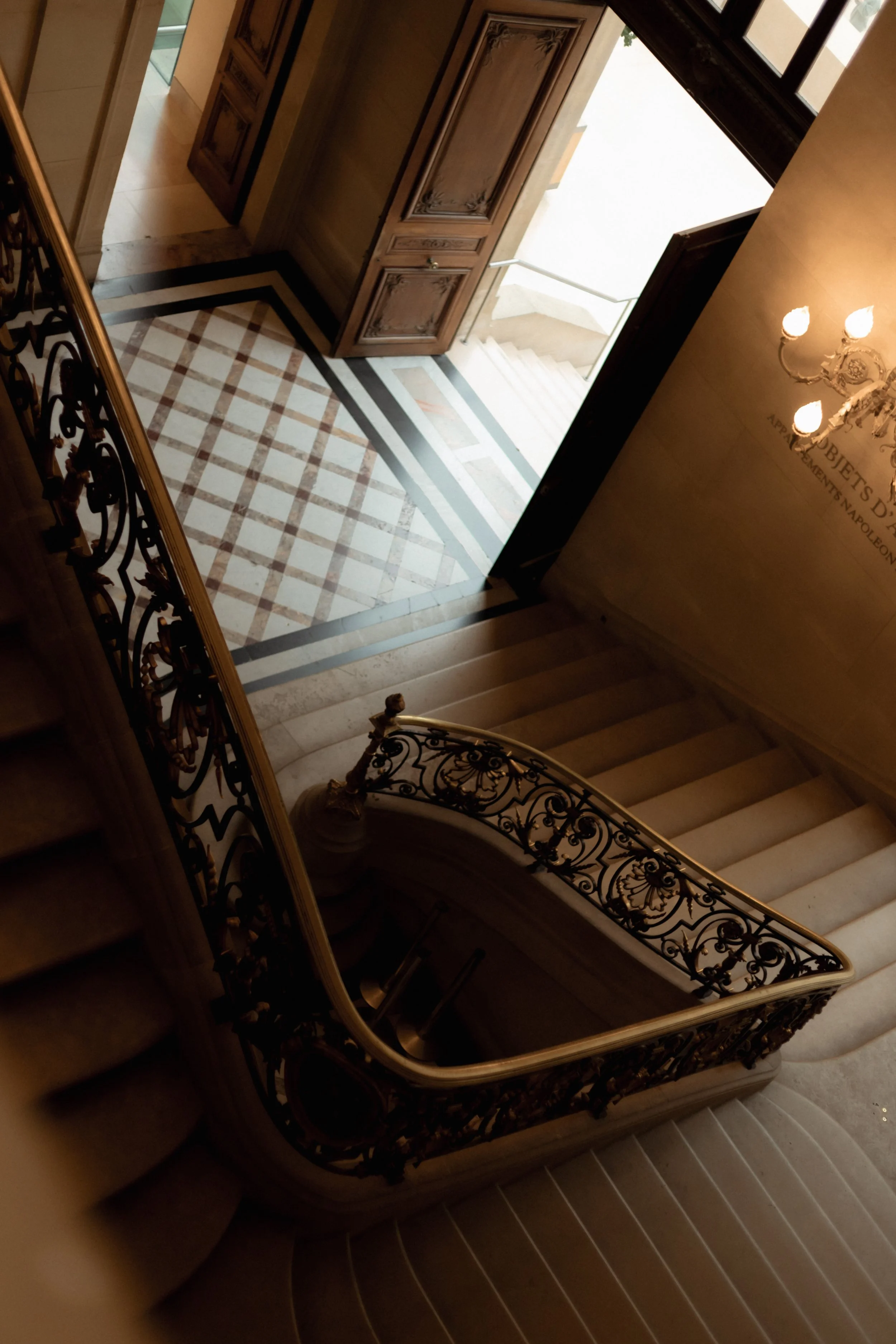 View down a grand stairway with ornate black and gold wrought iron railing, marble steps, and a decorative patterned marble floor at the bottom of the stairs, with wooden doors and a window