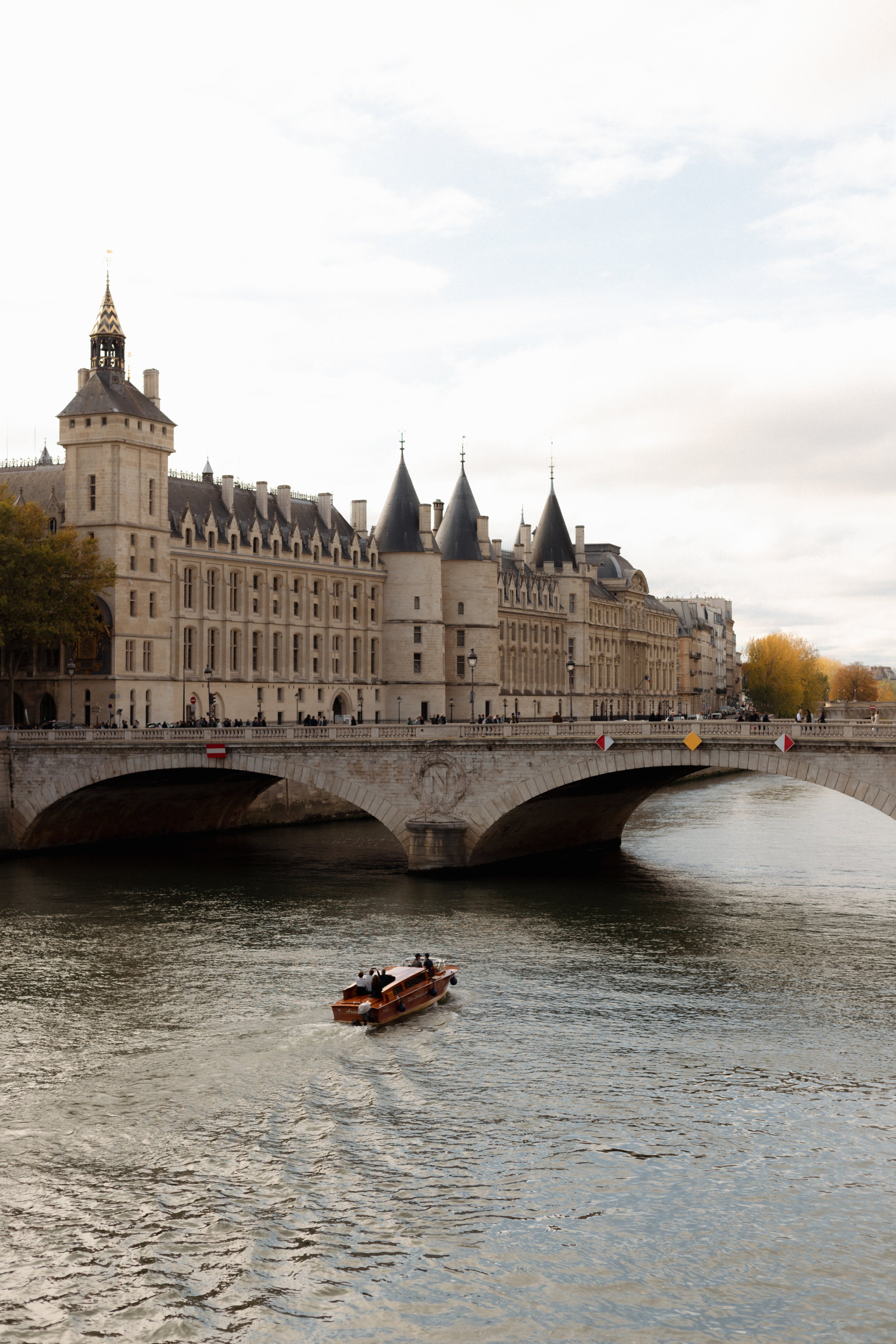 A boat with four people on the River Seine passing under a historic bridge, with the Conciergerie and other buildings in the background in Paris, France, during daytime.