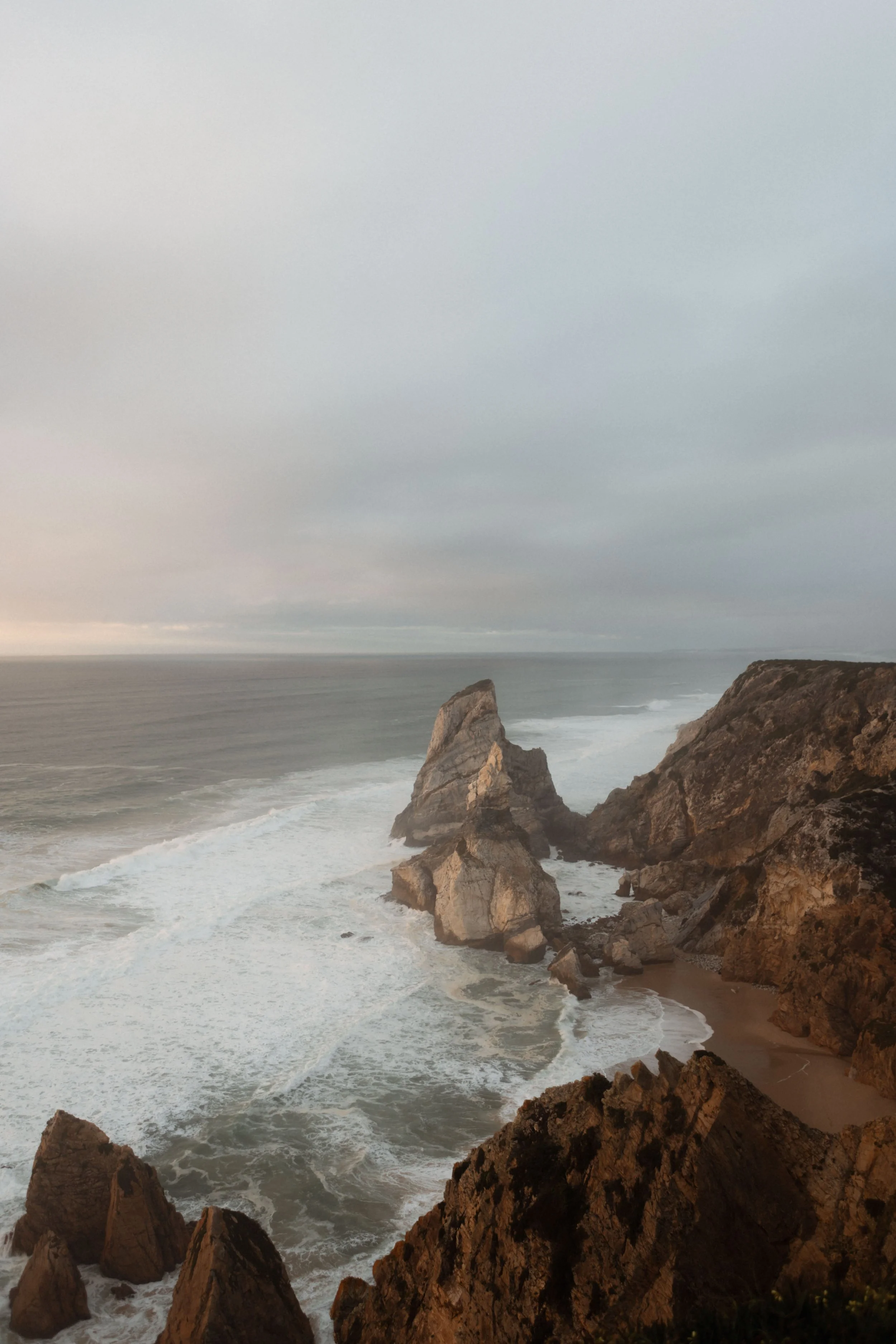 Coastal scene with rugged rocks jutting into the ocean, waves crashing against the rocks, under a cloudy sky.