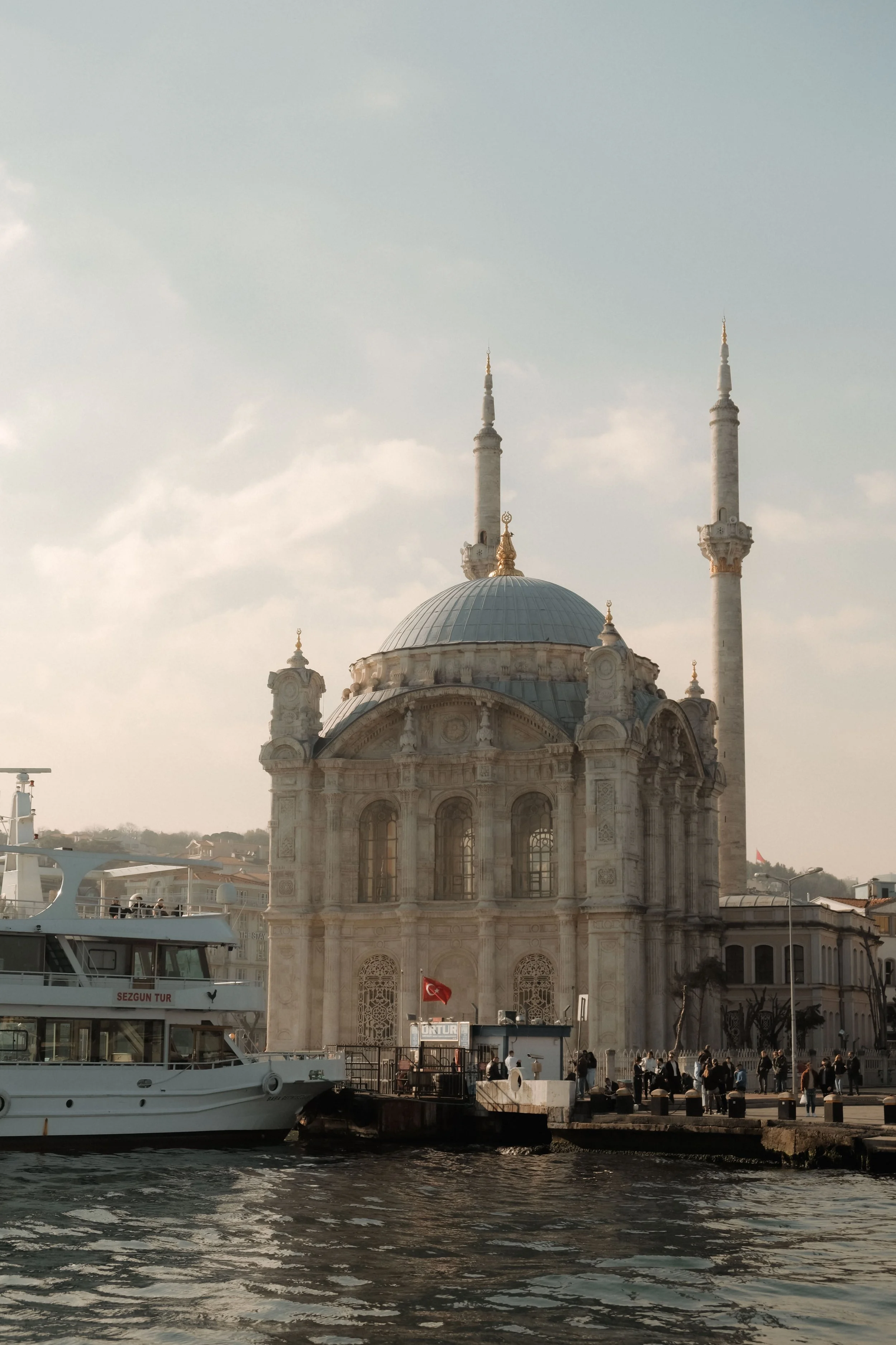 A historic mosque with domes and minarets next to a waterfront, with boats docked nearby and people walking along the promenade.
