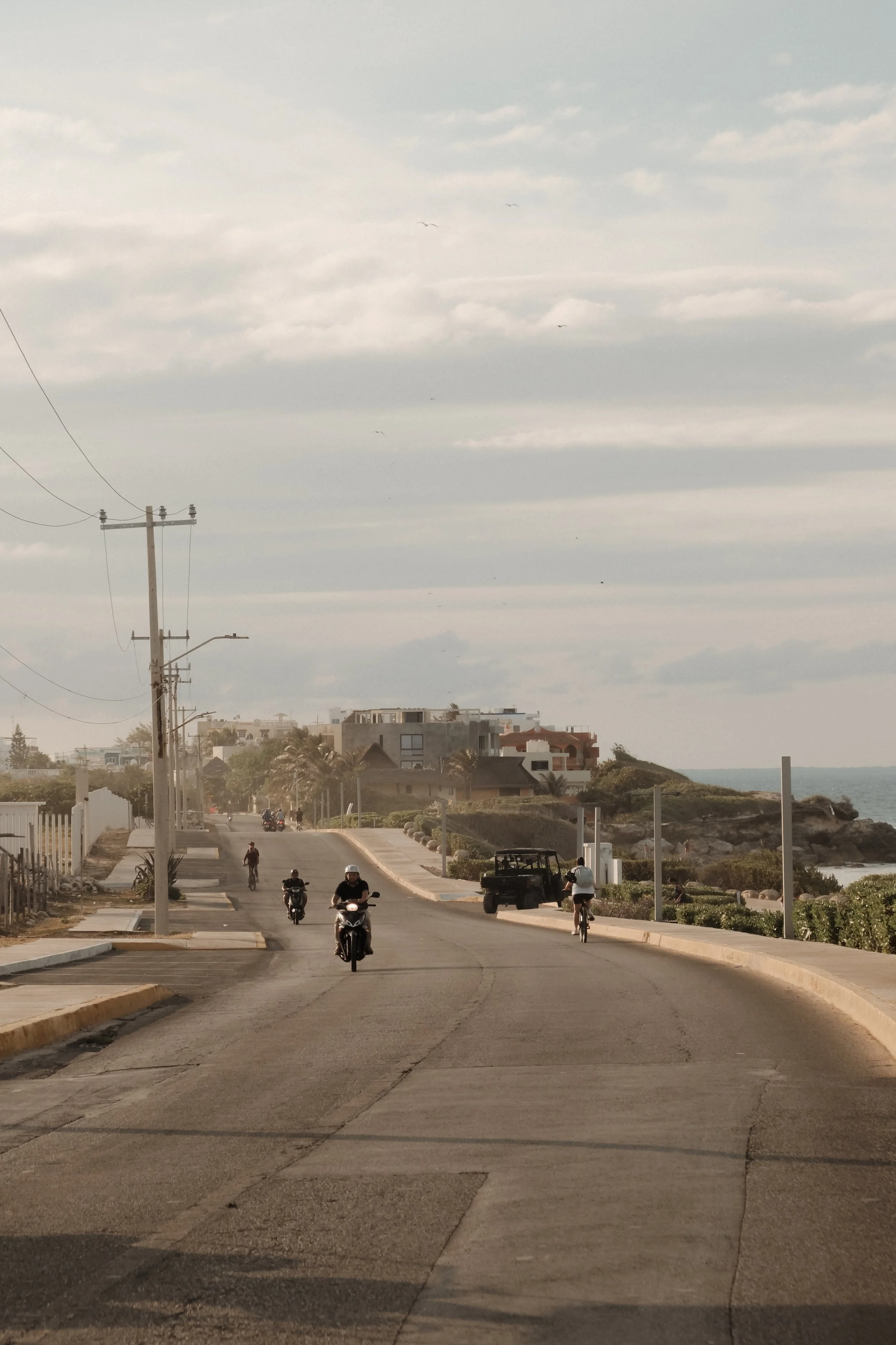 A street scene with people riding motorcycles and bicycles, a golf cart, and buildings in the background near the coast, with power lines and a cloudy sky above.