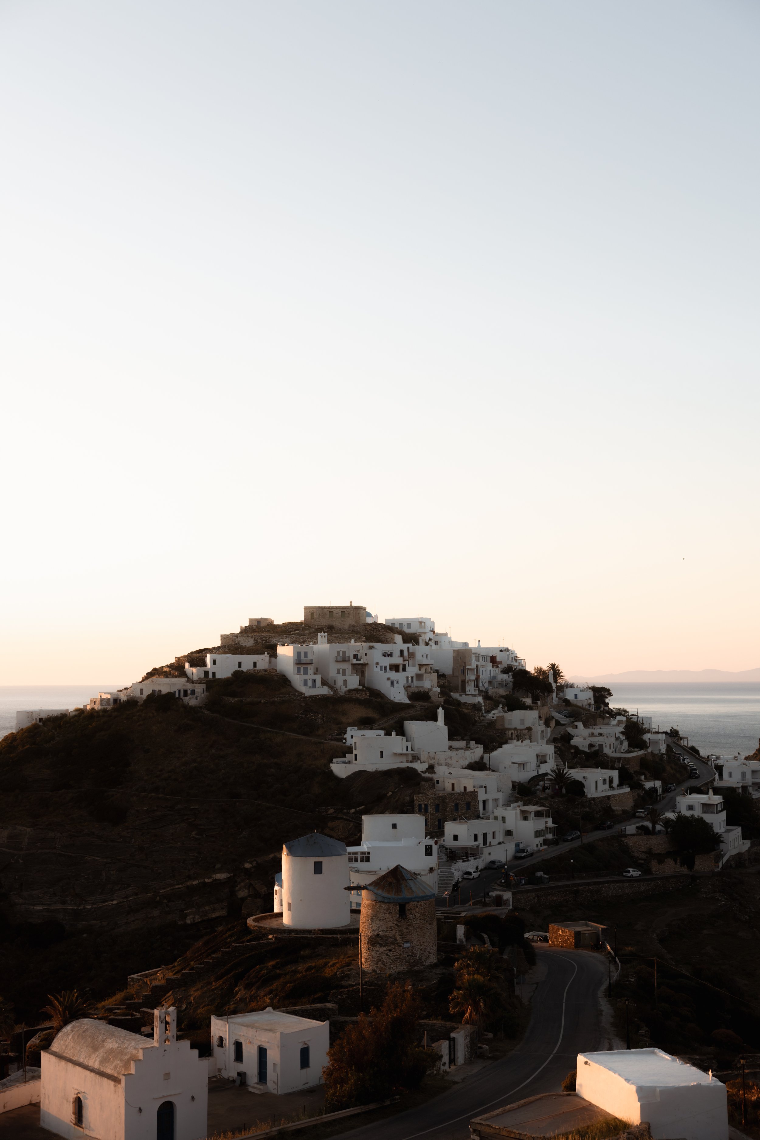 A hillside village with white buildings and windmills, overlooking a body of water, during sunset.