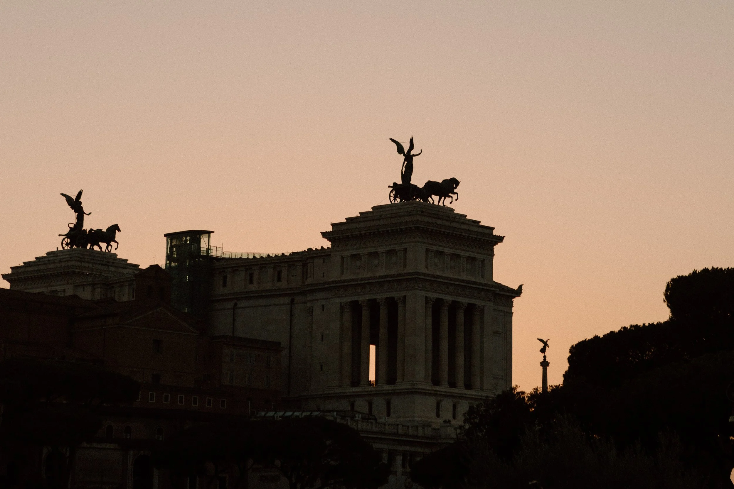 Silhouette of the Altare della Patria (Altar of the Fatherland) in Rome with sculptures of chariots and horses on top, against a pinkish sky during sunset.