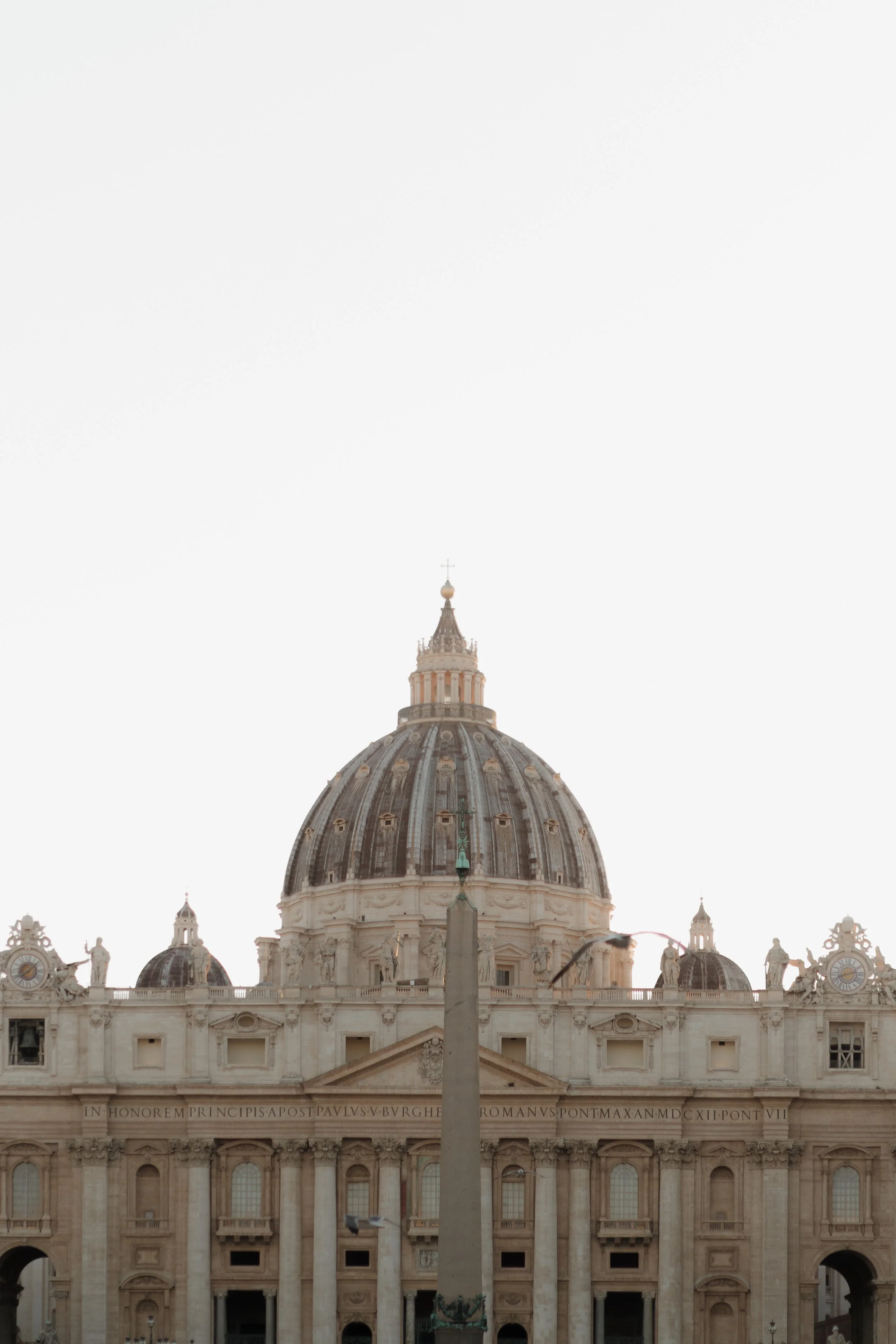 The dome of St. Peter's Basilica in Vatican City with an obelisk and statues in front, under a pale sky.