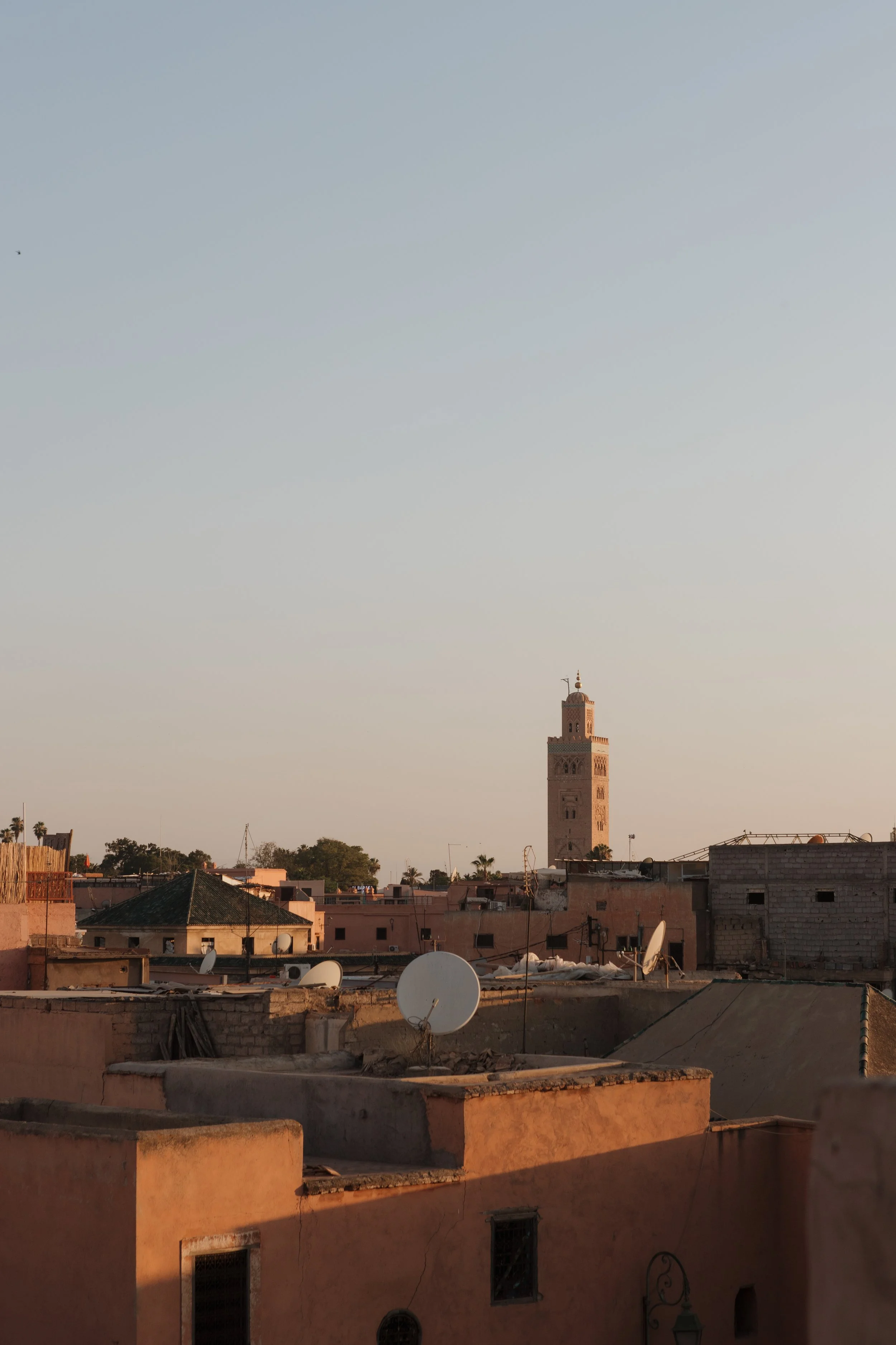 Cityscape at sunset featuring a tall mosque with a minaret, surrounded by flat-roofed buildings and satellite dishes, under a clear sky.