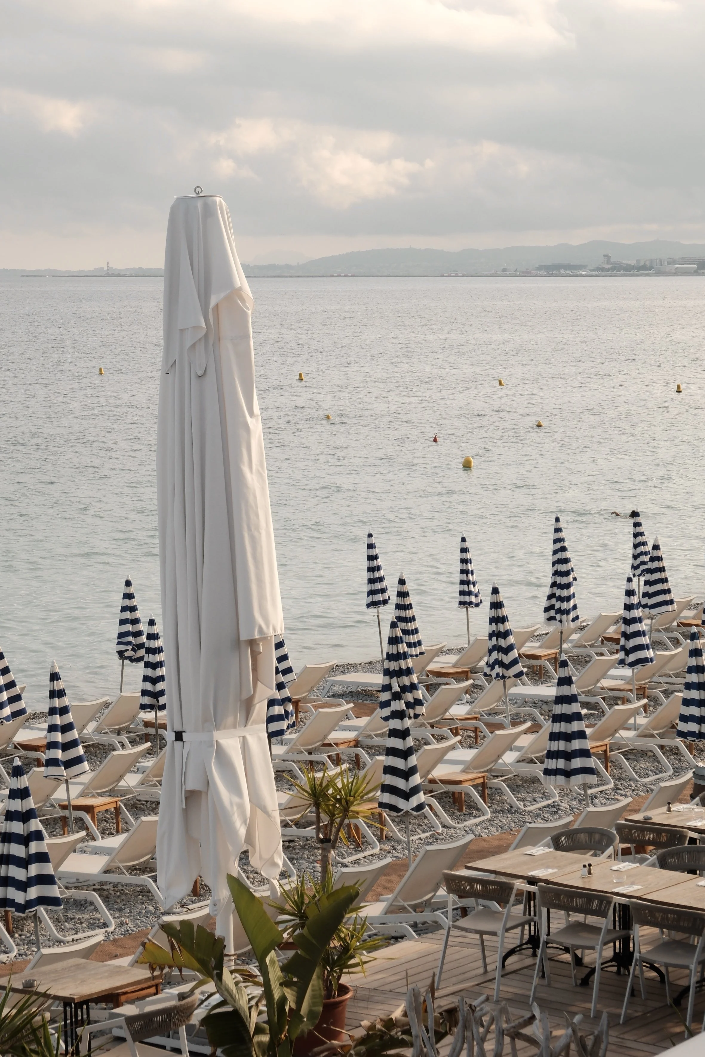 Empty beach restaurant with white and black striped umbrellas, white chairs, and tables overlooking the water and cloudy sky.