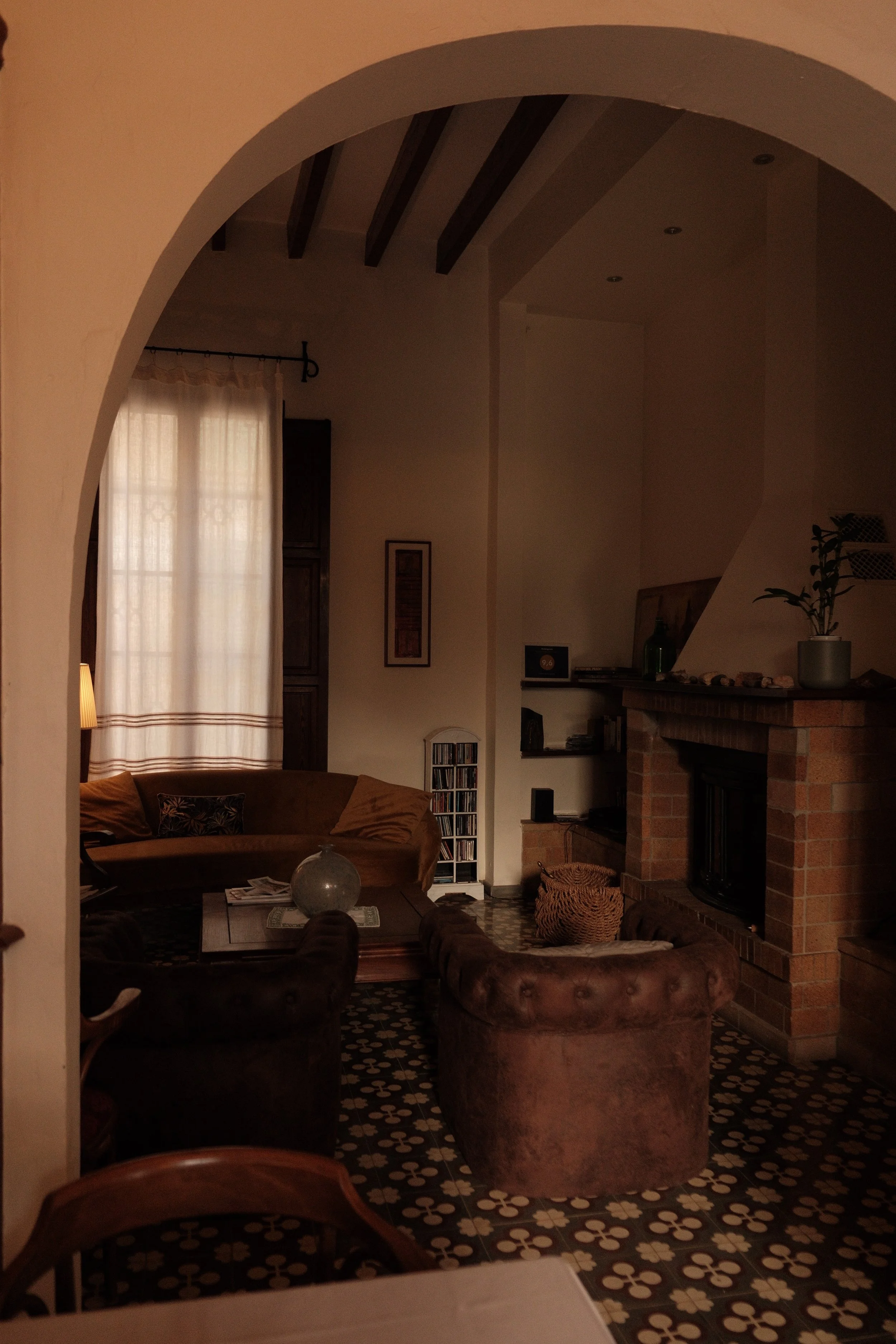 Living room with a high ceiling, arched doorway, and brick fireplace. Contains a brown velvet armchair, a coffee table with a glass orb, and a patterned tile floor. Light-colored curtains cover a window.