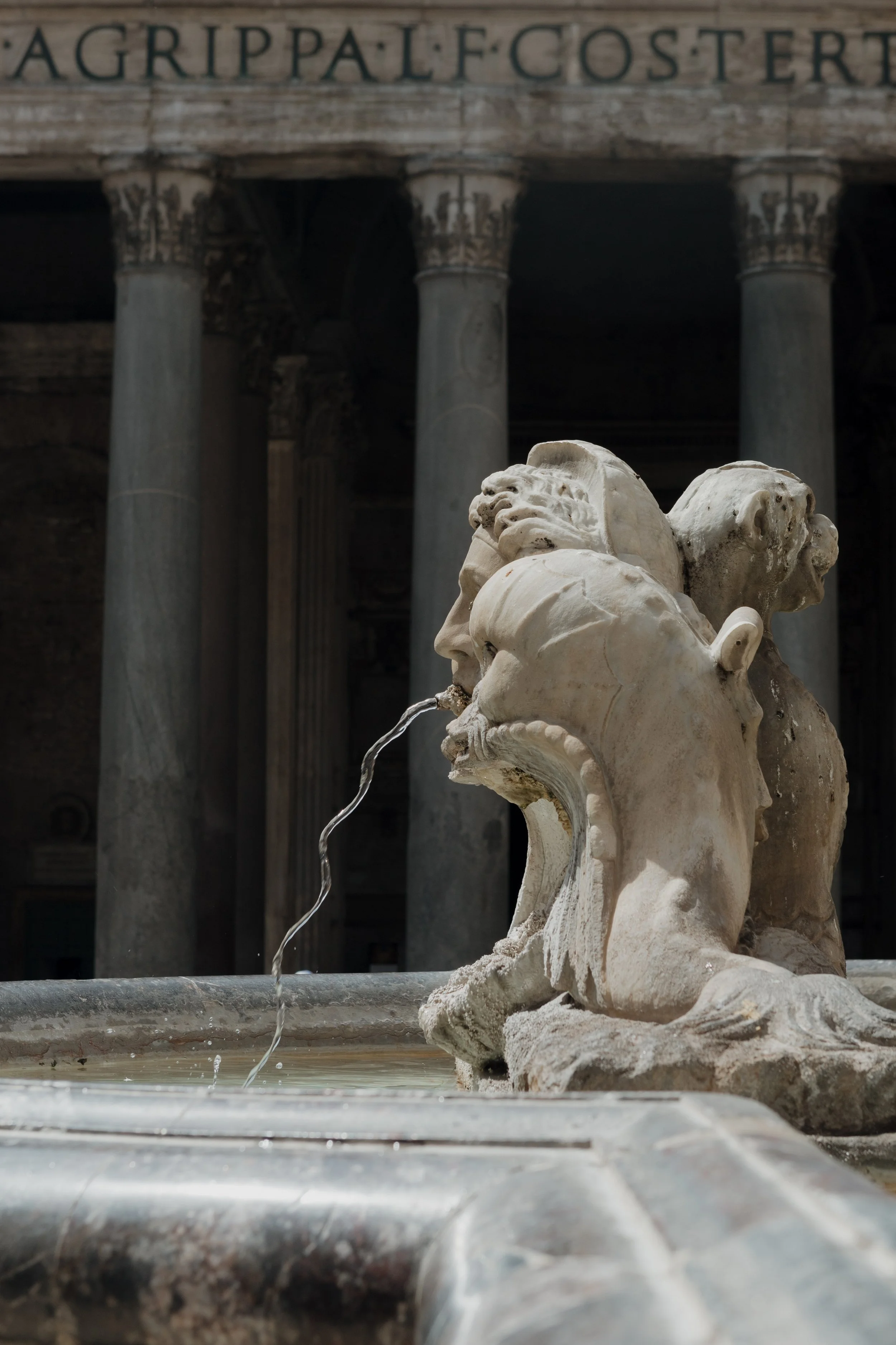A marble sculpture of a lion's head water fountain outside ancient Roman ruins with large stone columns in the background.