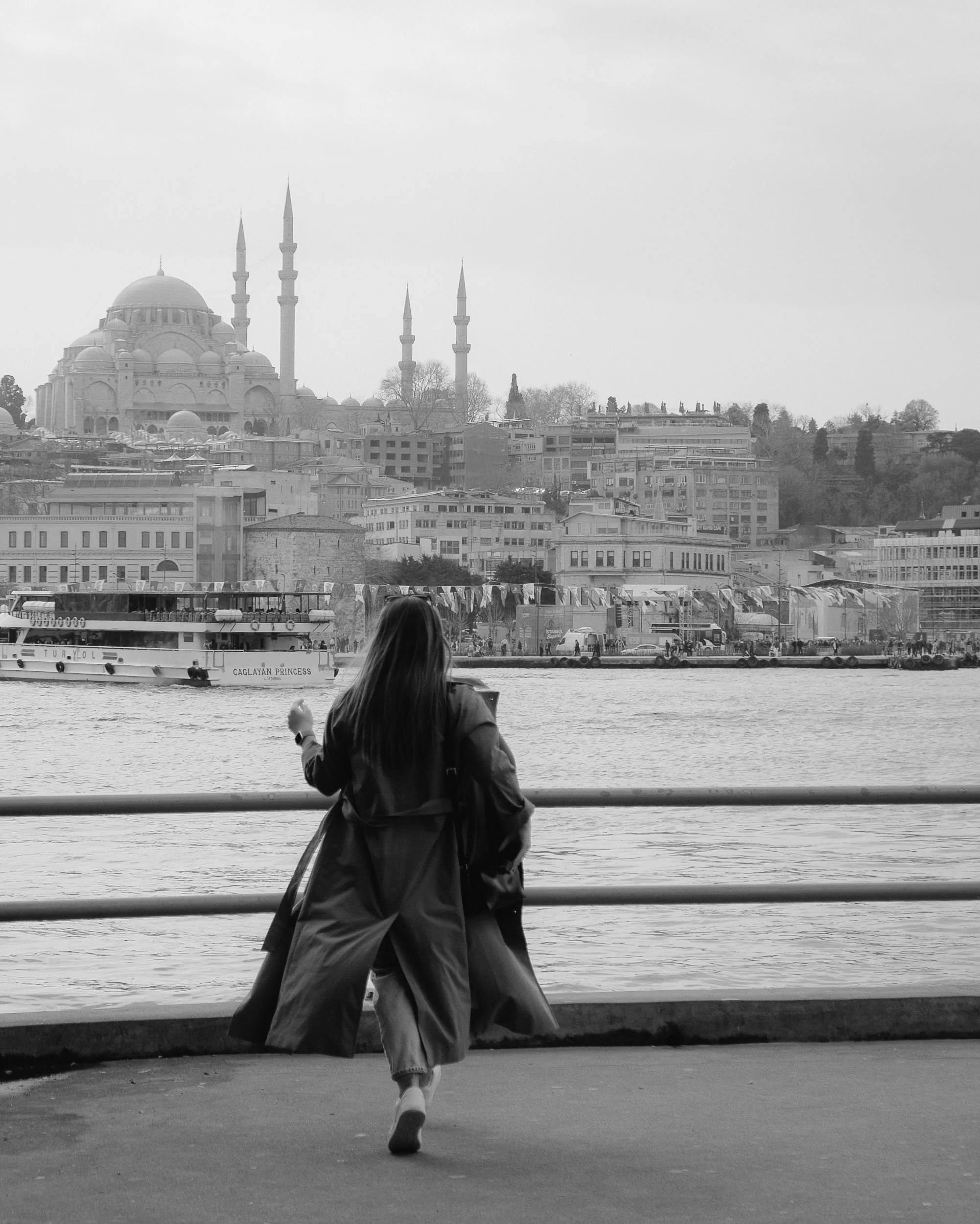 A woman with long dark hair and a coat leaning against a metal railing by the water, overlooking a cityscape with a large mosque with multiple domes and minarets in the distance, black and white photo.