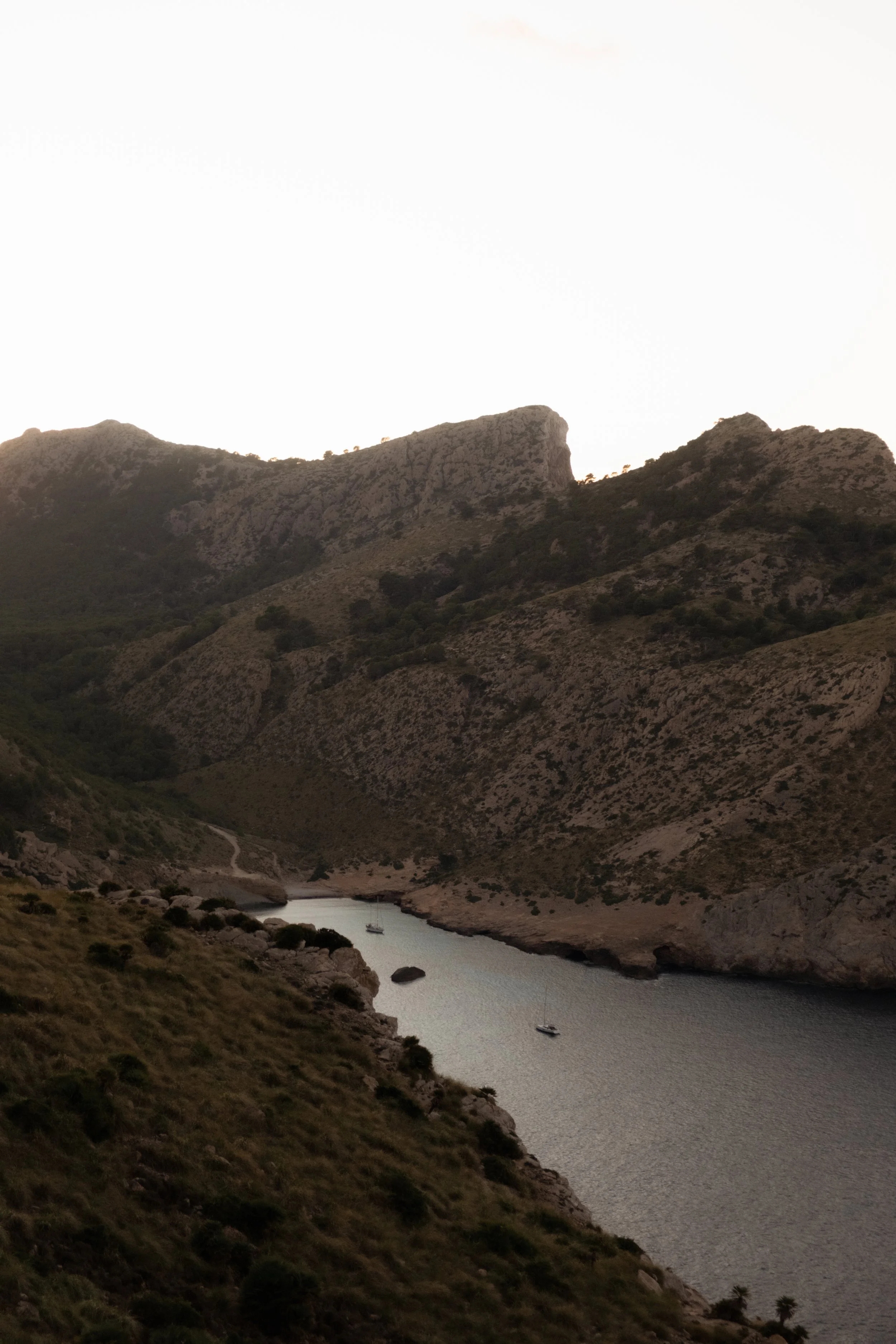 Landscape of a river flowing through mountainous terrain with rocky hills, sparse vegetation, and a sailboat on the water, under an overcast sky.