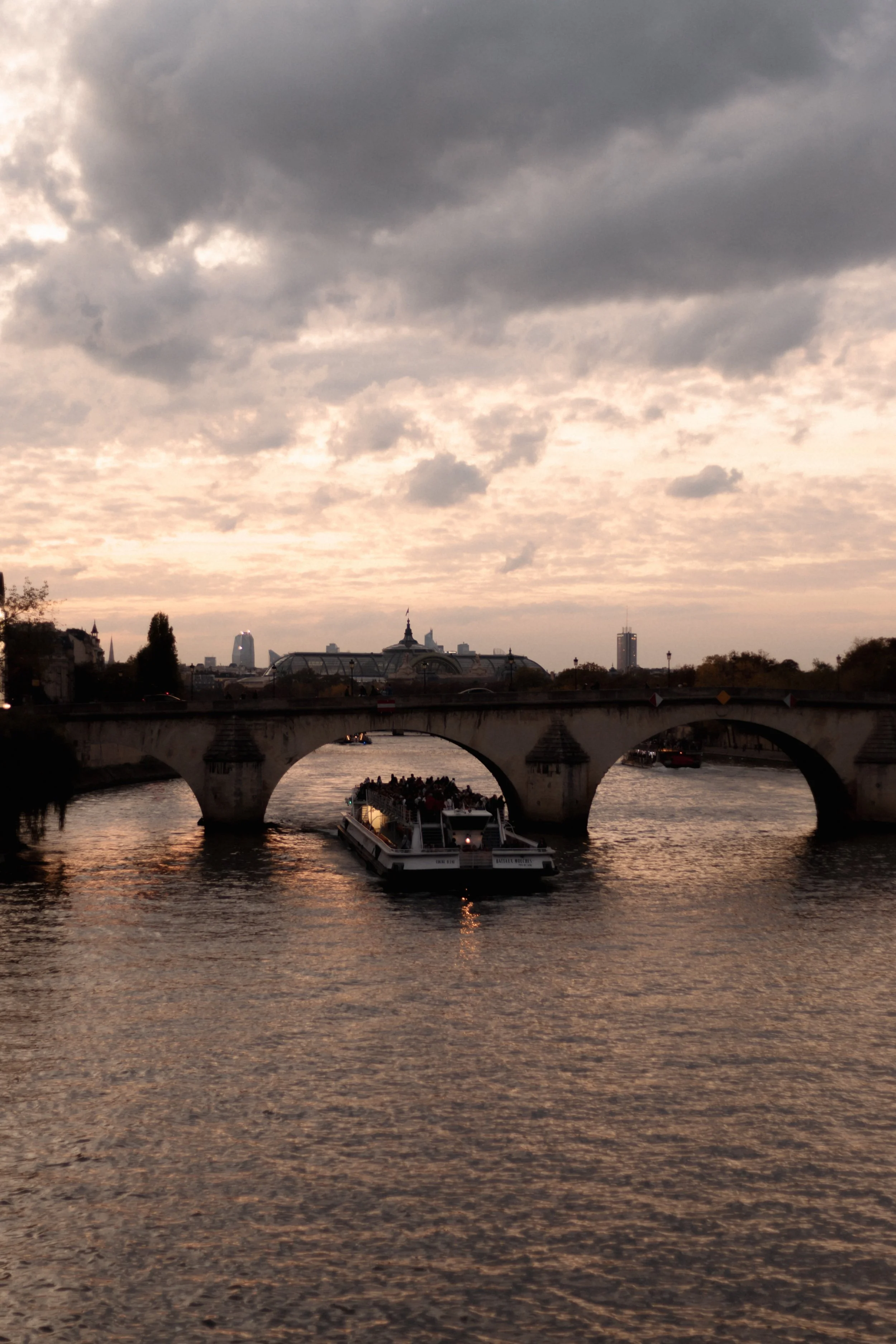 A boat ferry floating under a stone bridge on the River Seine in Paris during dusk, with a cloudy sky overhead.