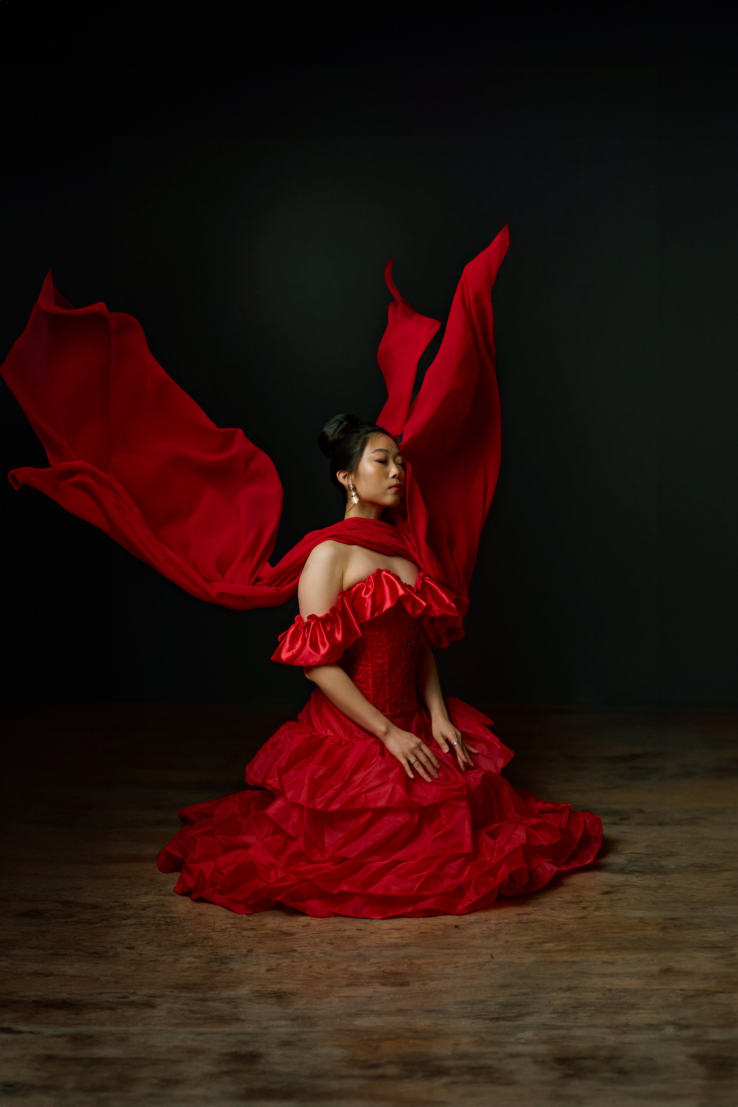 A woman in a red flowing dress with a corset-style bodice, sitting on a wooden floor with her eyes closed and hands on her lap, against a black background.